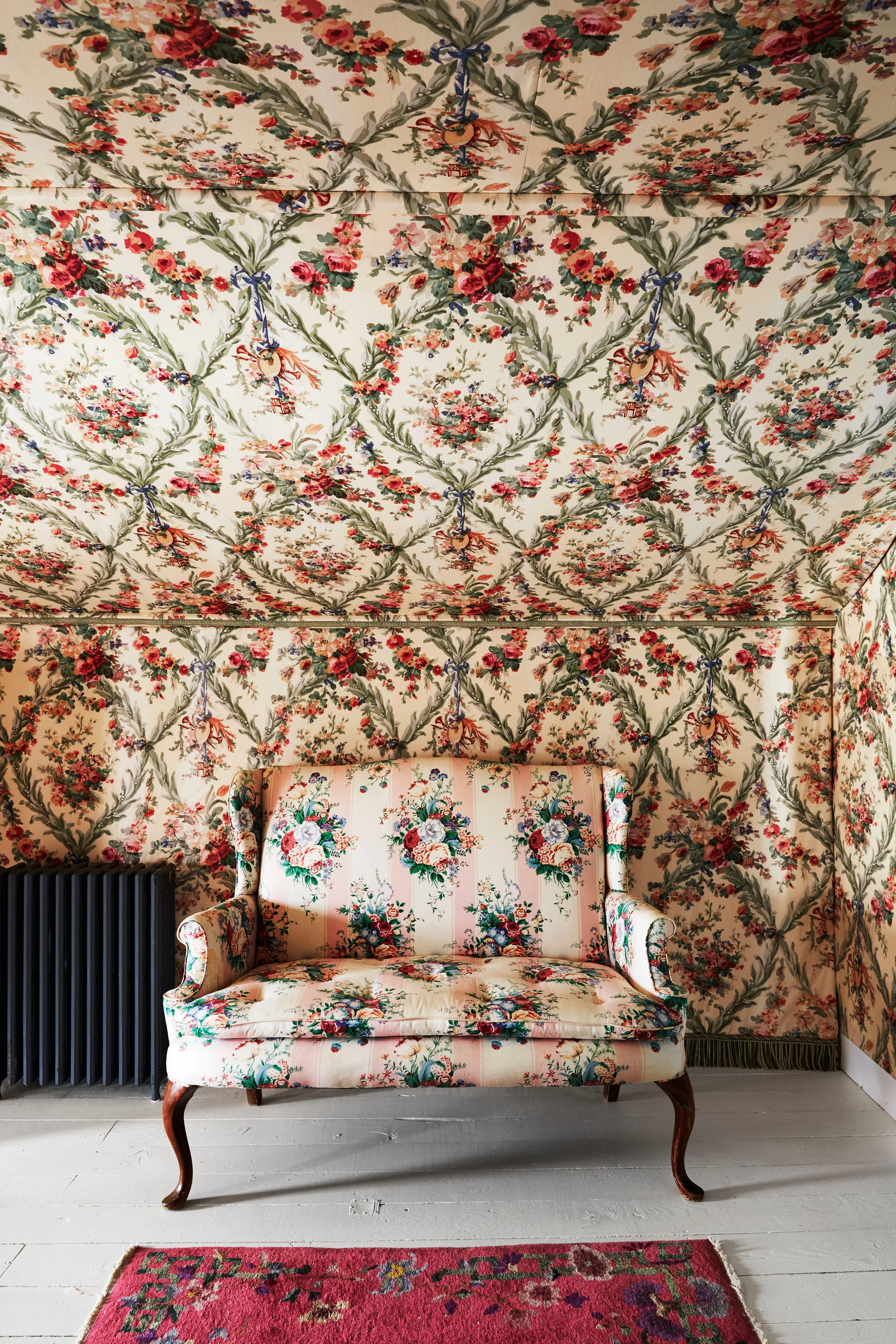 A vintage floral settee sits against walls and a ceiling covered in matching red and green floral wallpaper, on white painted wooden floors.