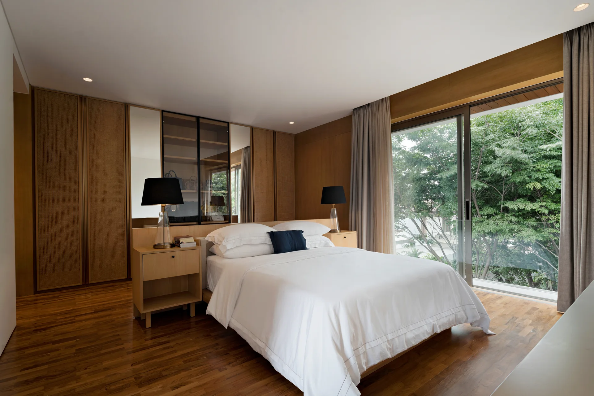 A contemporary bedroom featuring a bed with white linens, built-in wood and cane cabinets, matching nightstands, and a large window looking out to trees.