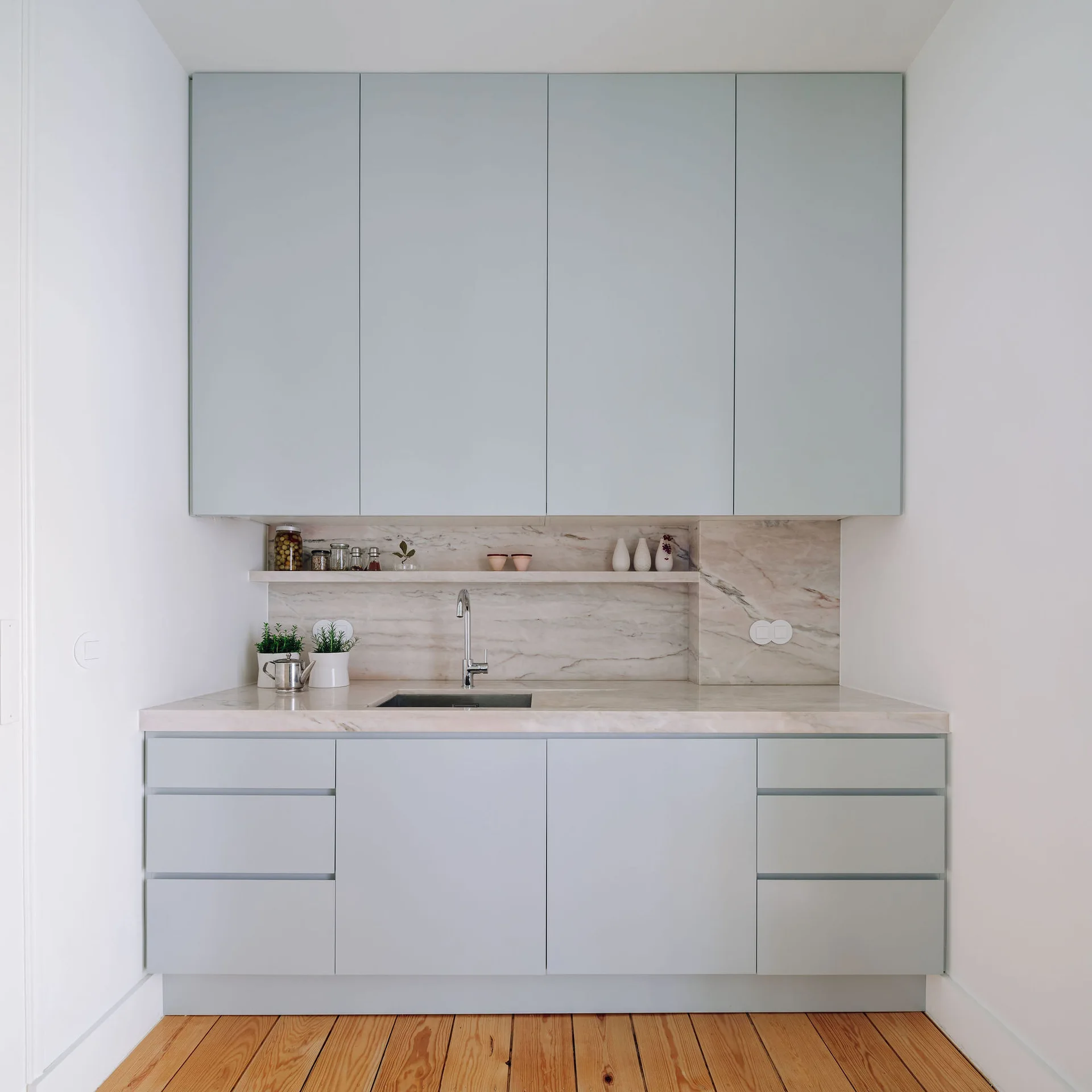 A minimalist kitchen niche with pale blue handleless cabinets, a veined marble countertop, and matching backsplash with a slim floating shelf.