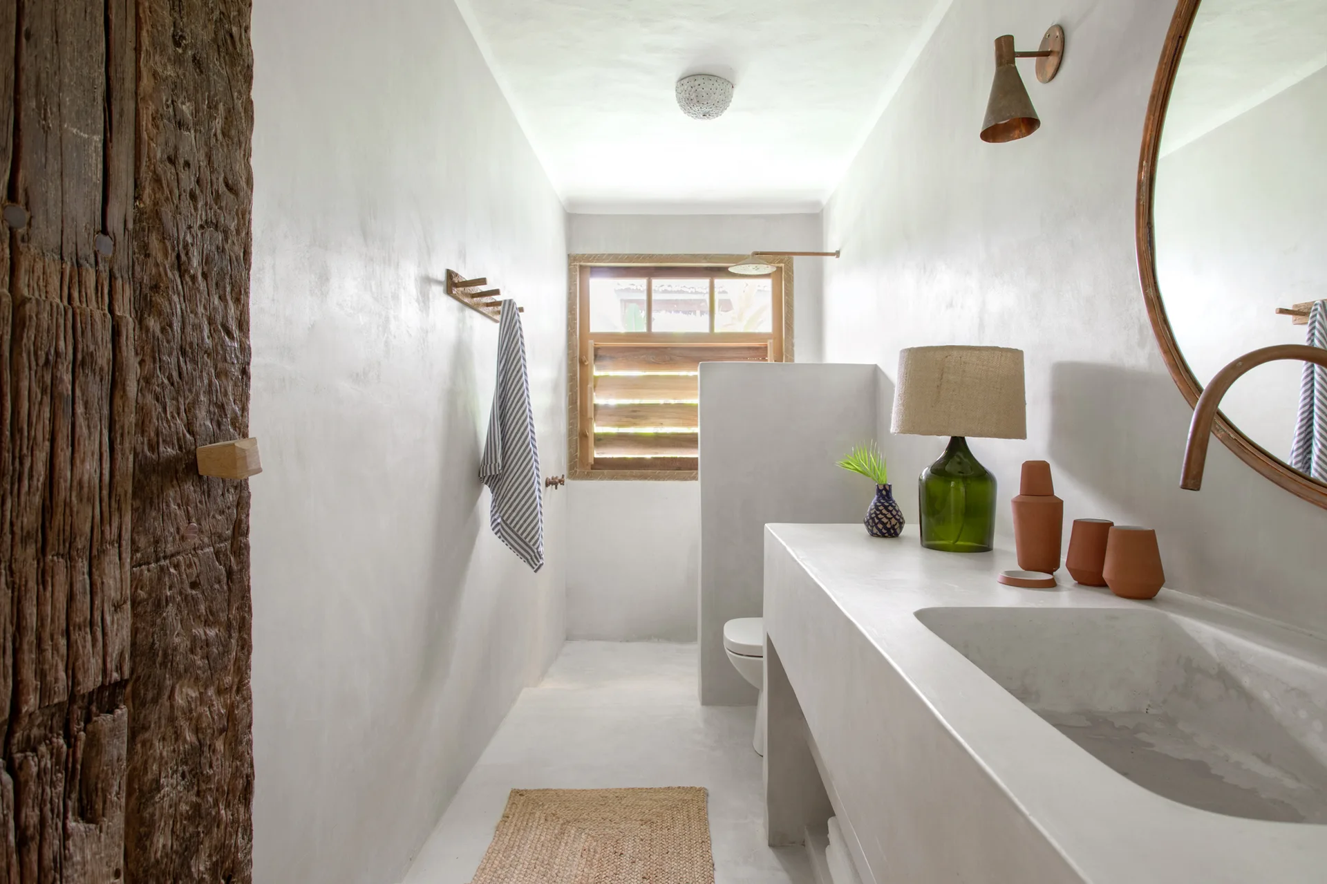 A narrow minimalist bathroom with an integrated concrete vanity, plaster-finish walls, a wooden louvered window, and a rustic timber pillar.