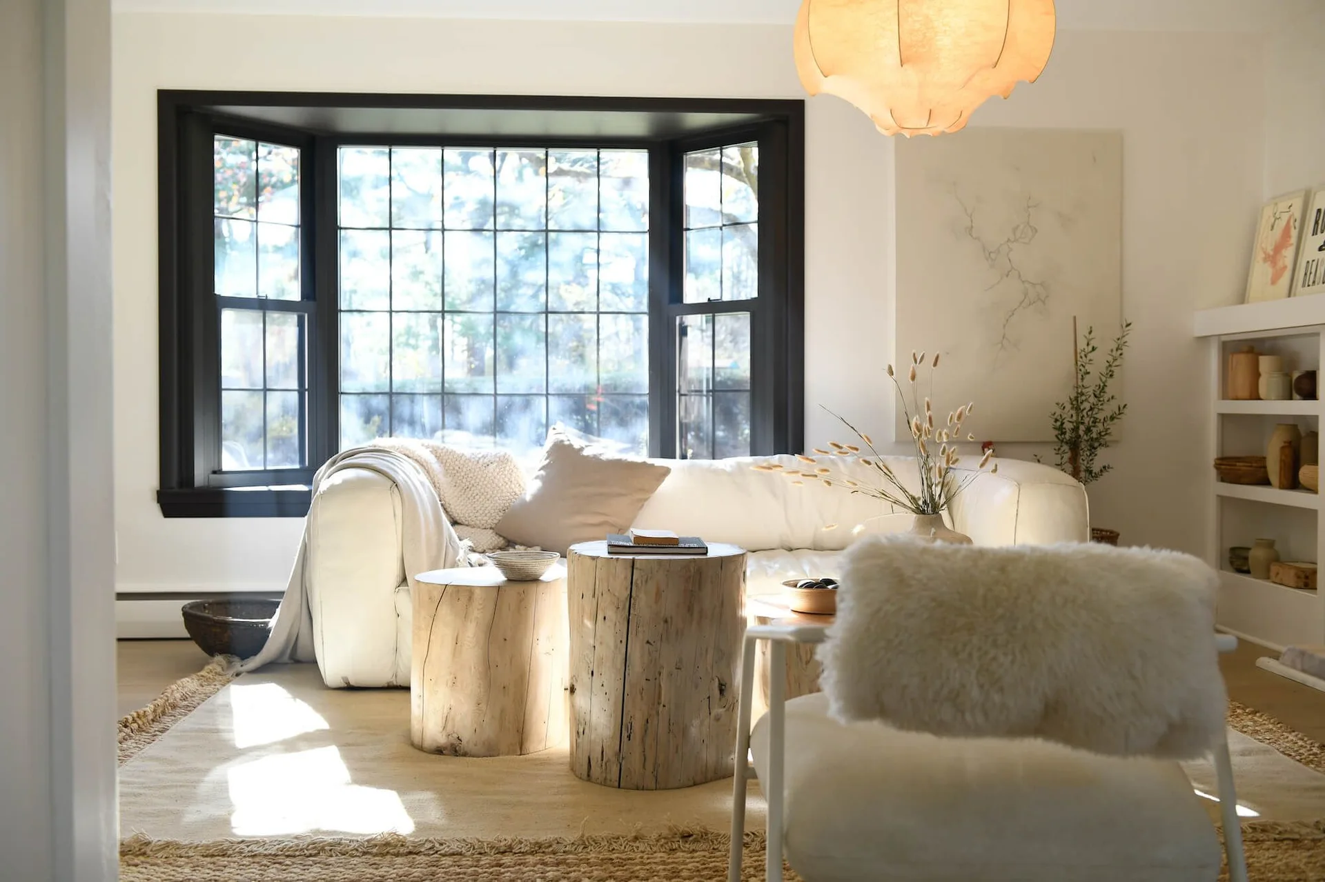 A bright living room with a white sofa, raw wood stump coffee tables, a textured rug, and a bay window featuring black frames.