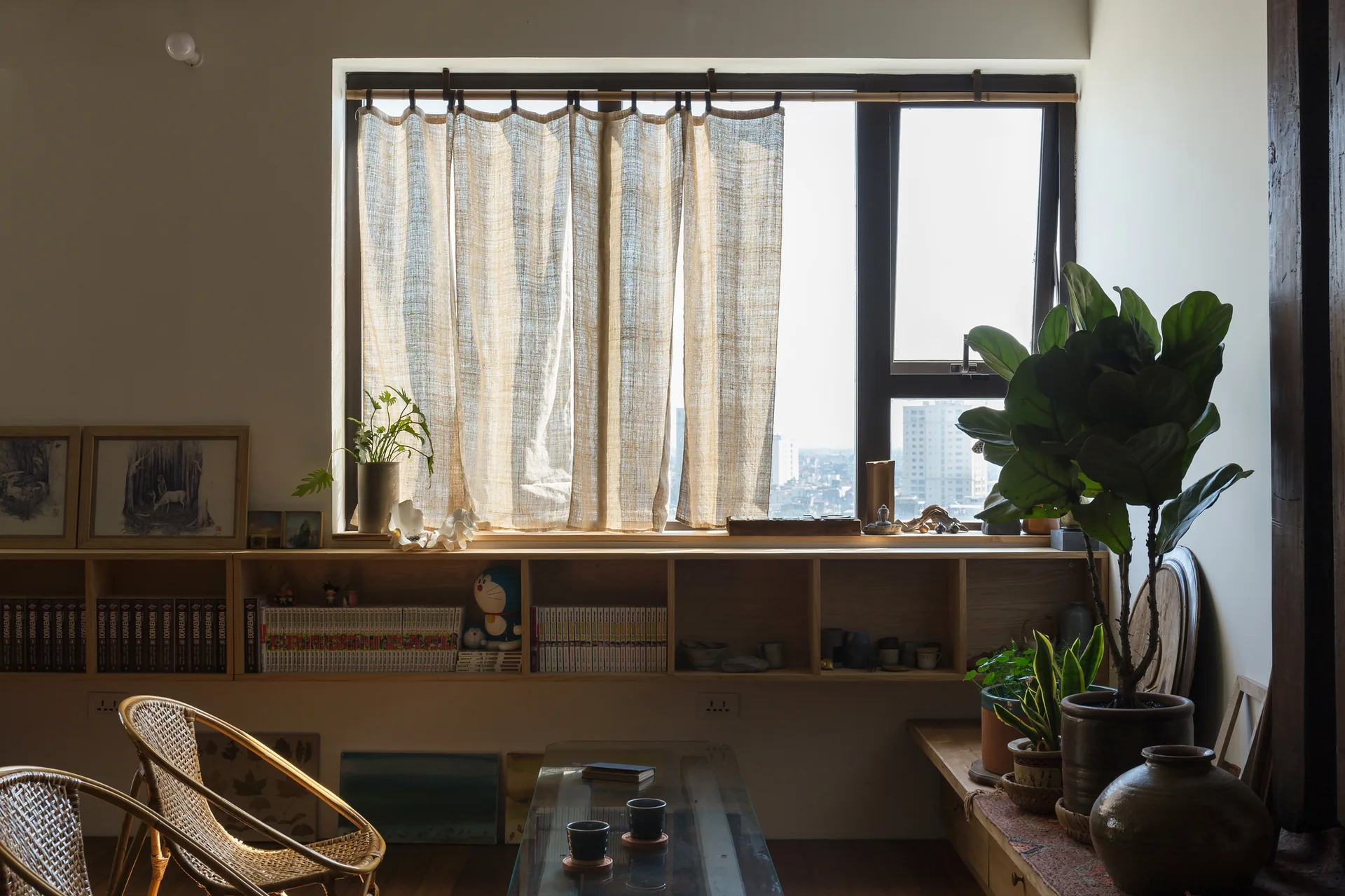 A sunlit living room features a low wooden bookshelf built under a large window with sheer linen curtains, rattan chairs, and potted plants.