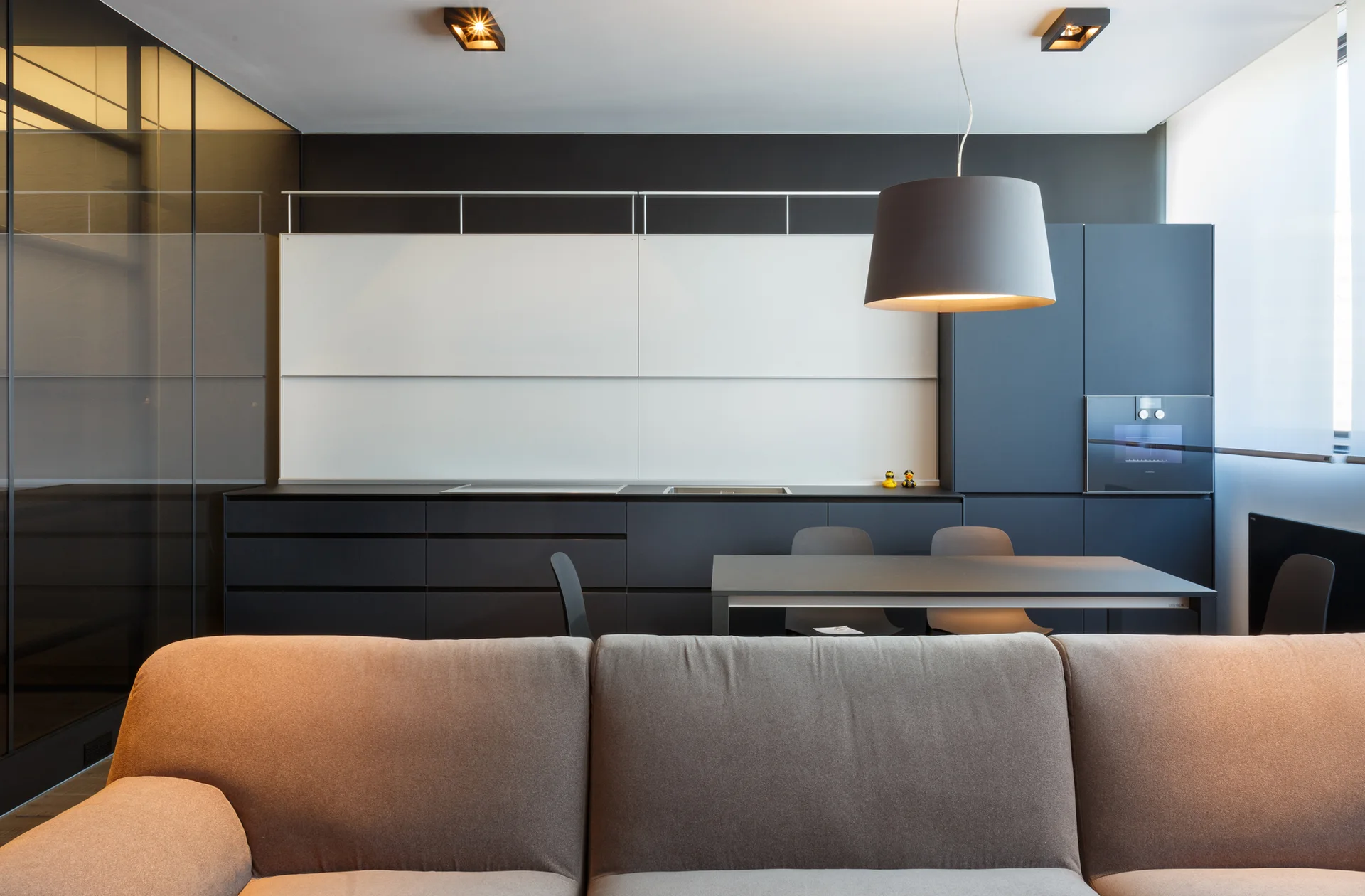 A modern open-plan kitchen features dark grey cabinetry, a dining table under a pendant light, and a tan sofa in the foreground.
