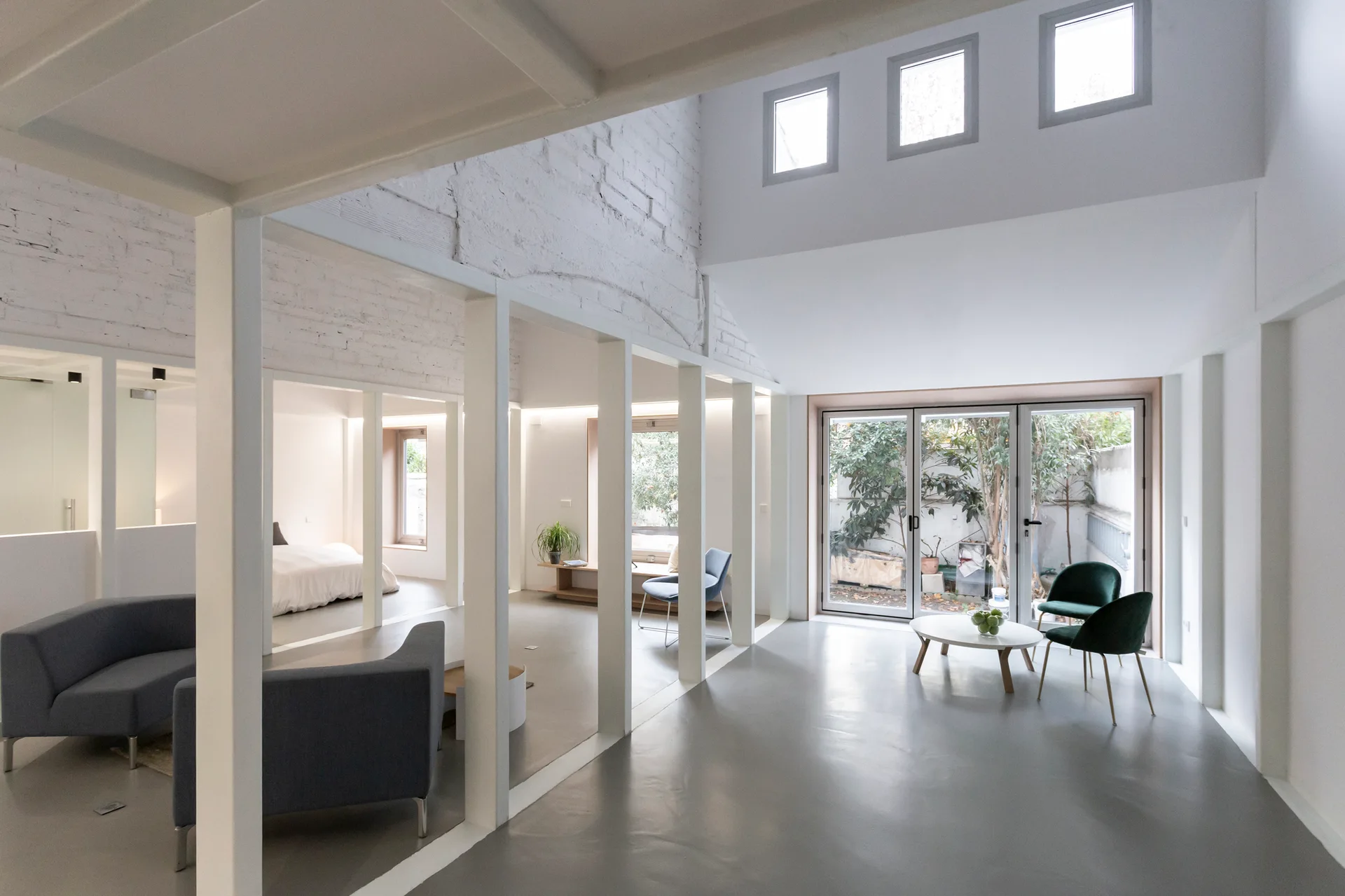 An open-plan loft features white structural columns, a smooth gray floor, white-painted brick walls, and modern seating areas near glass doors.