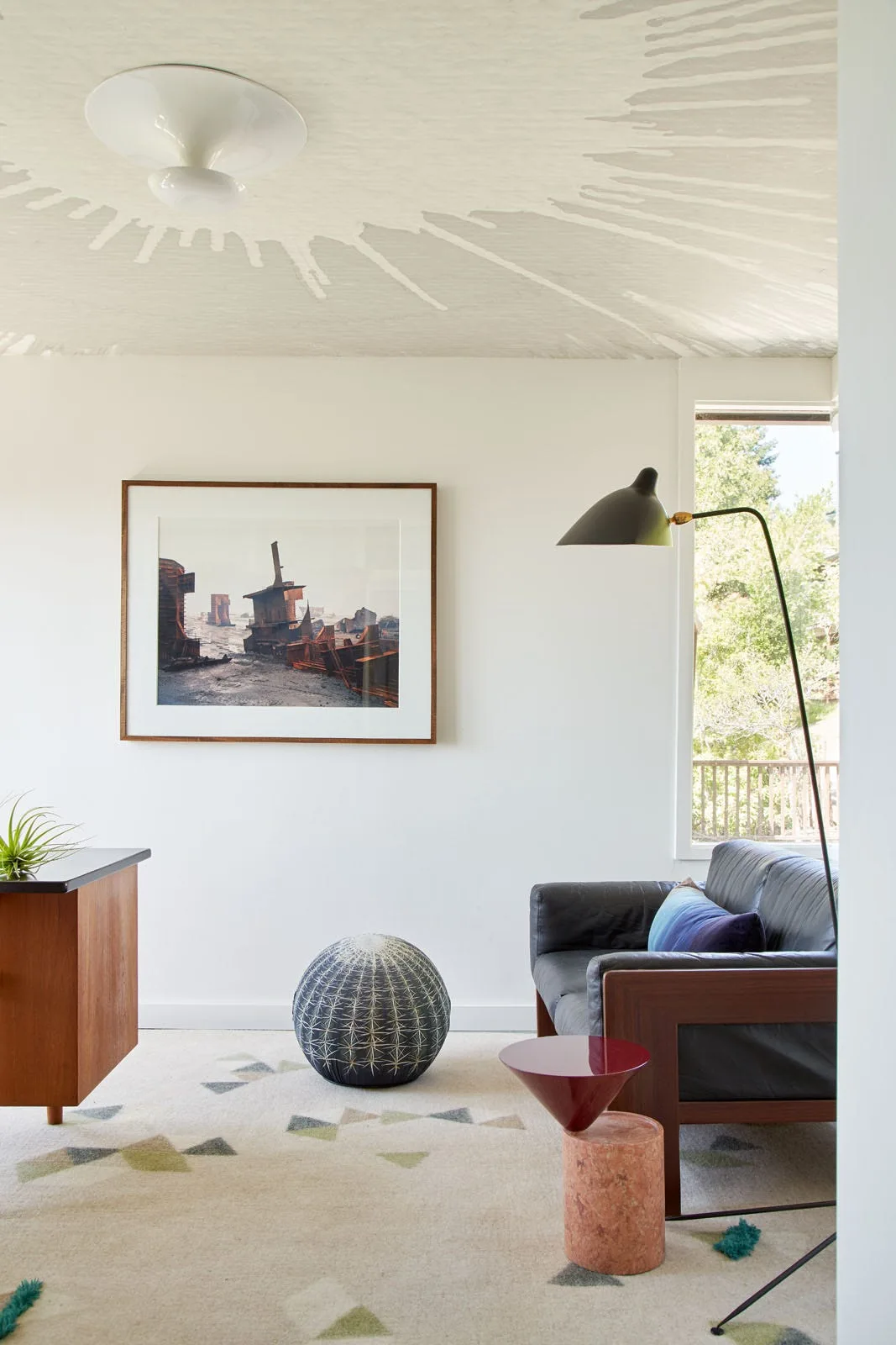 A modern living room features a leather sofa, a cactus-patterned pouf, a black floor lamp, and a large abstract mural painted on the ceiling.