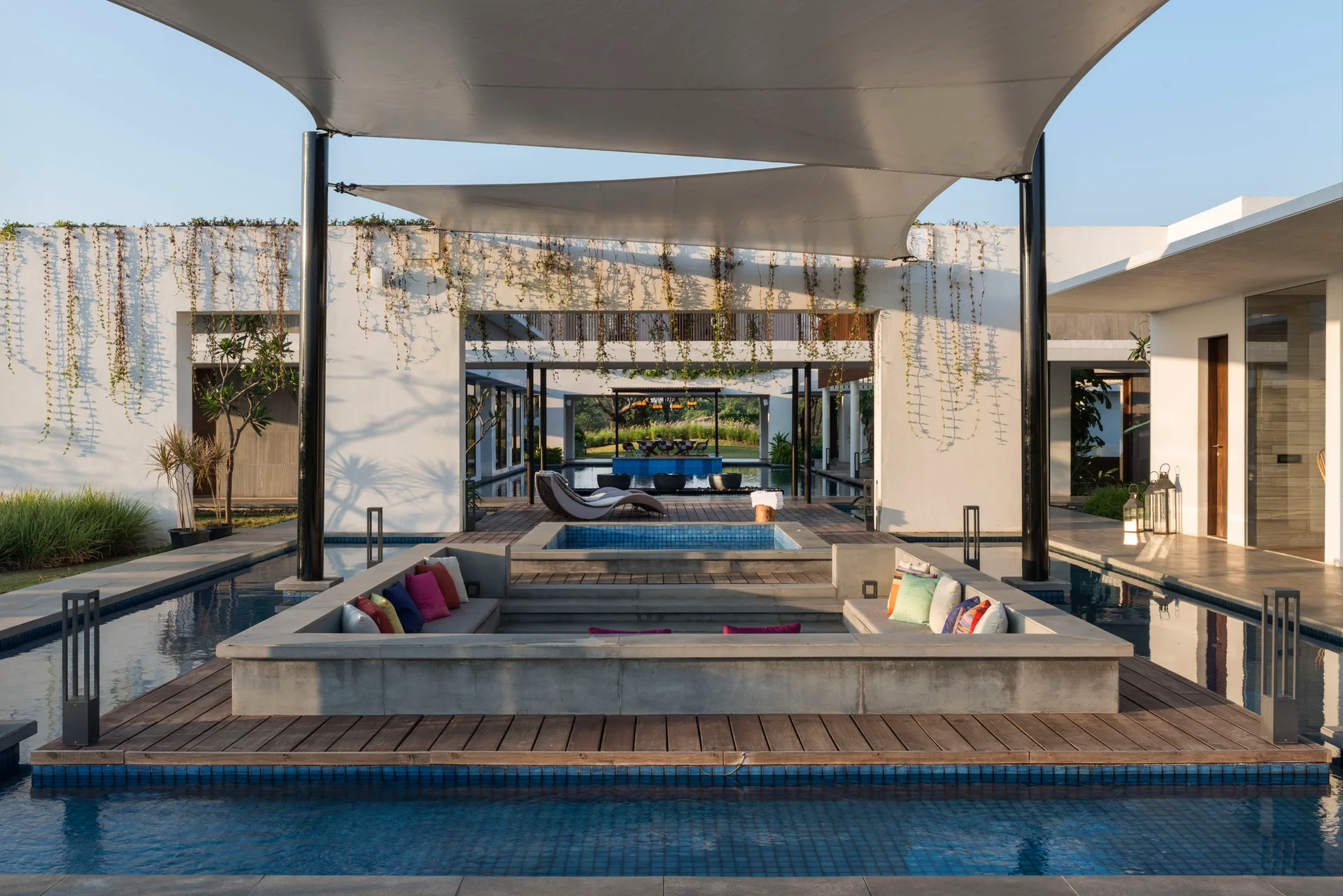 A modern outdoor sunken concrete seating area with colorful pillows is surrounded by a pool, wood decking, and a white overhead canopy.