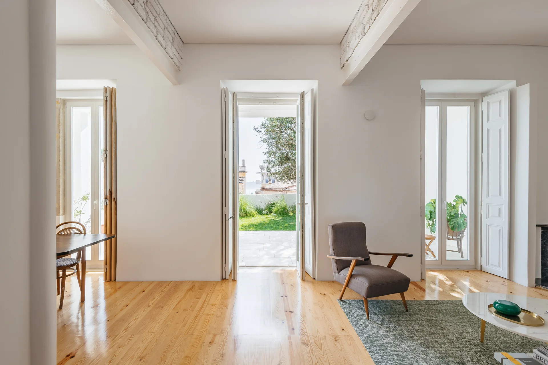 A bright living room with polished light hardwood floors, a gray armchair, and tall French doors that open to a green terrace.