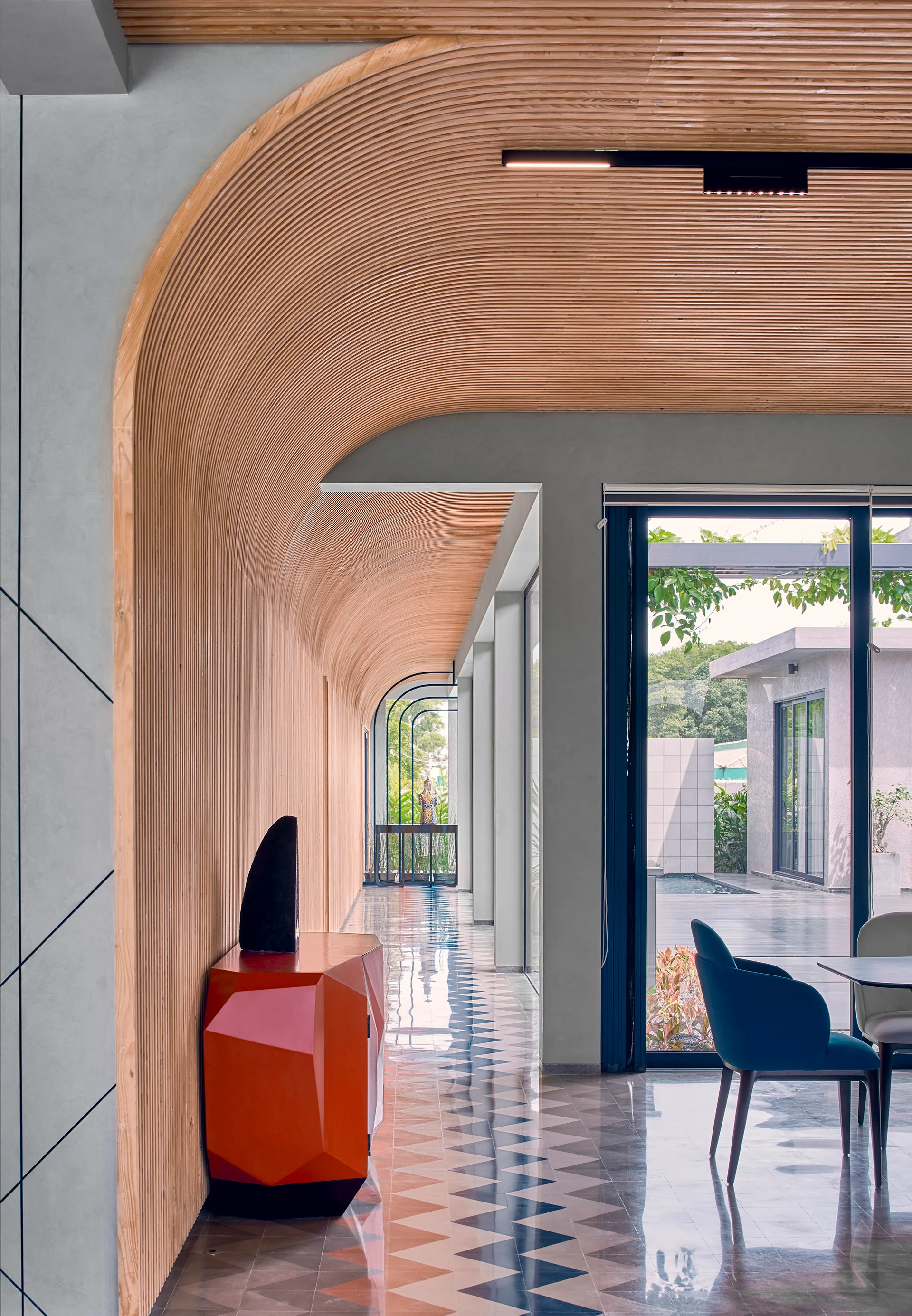 A long hallway features a curved slatted wood ceiling, geometric patterned floor tiles, and an orange console table next to large glass doors.