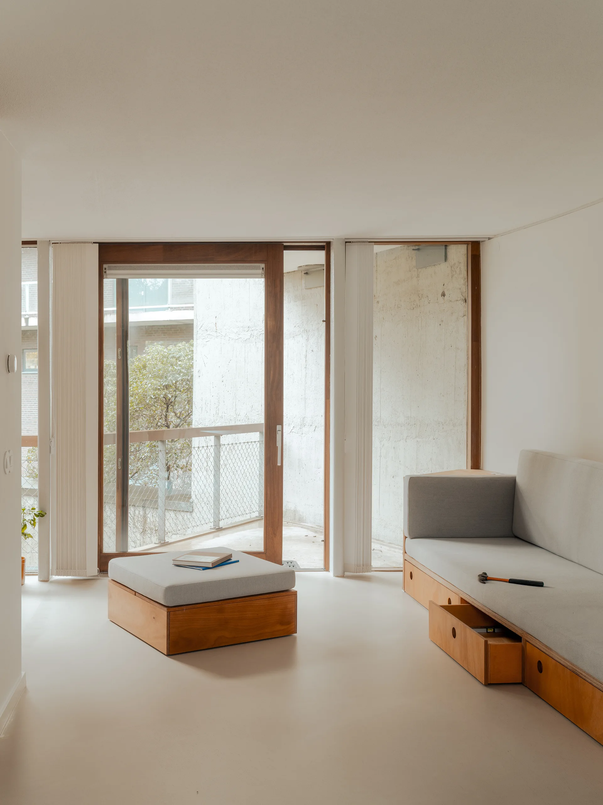 A minimalist living room features a natural wood sofa with built-in under-seat drawers, a matching low ottoman, and sliding doors opening to a balcony.