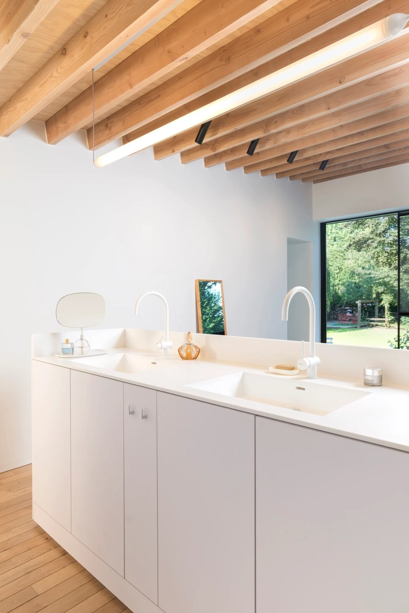 A minimalist white double-sink bathroom vanity with two white gooseneck faucets sits beneath an exposed light wood joist ceiling.