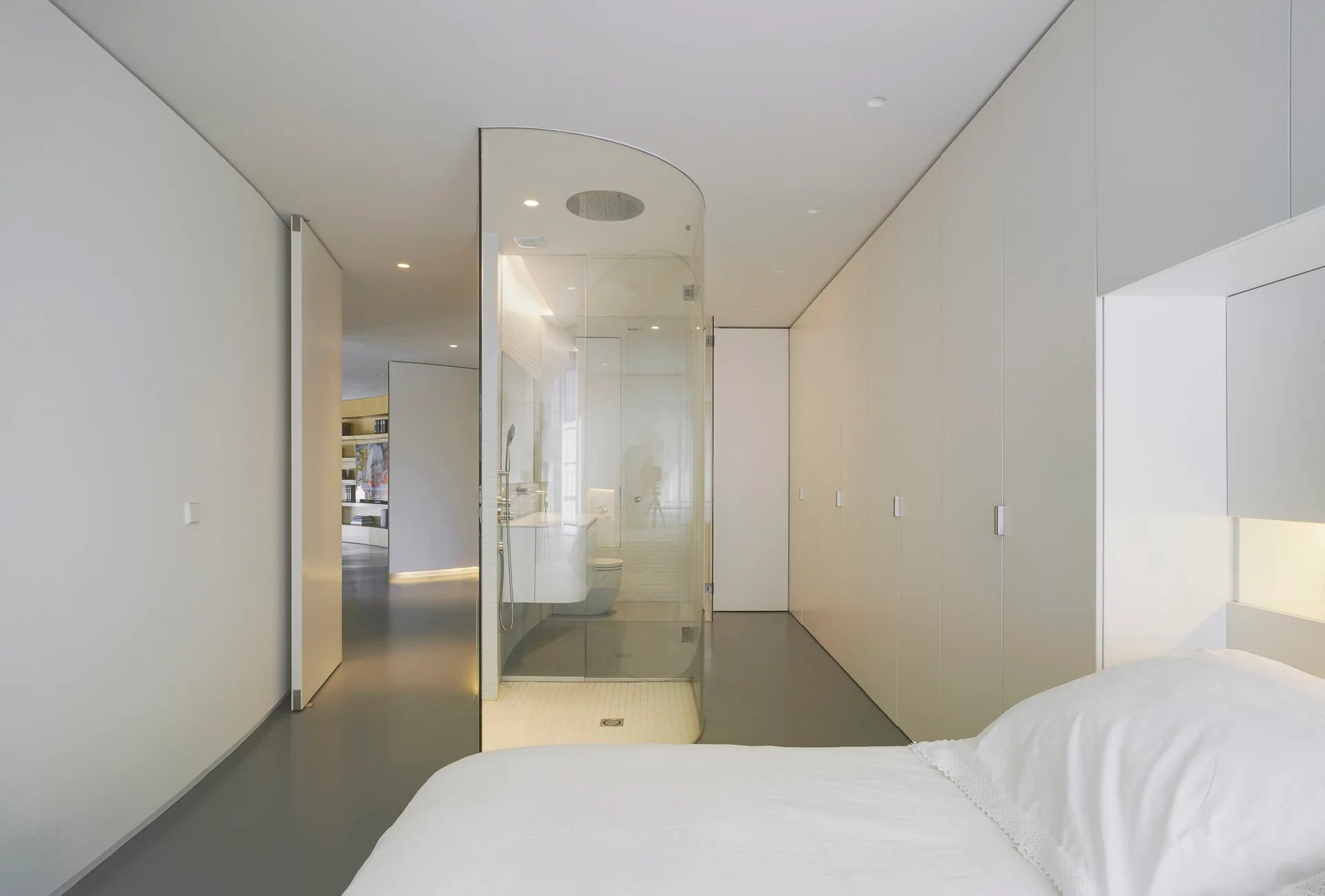 A minimalist bedroom with a curved glass shower unit, built-in wardrobes, and white bedding in the foreground.