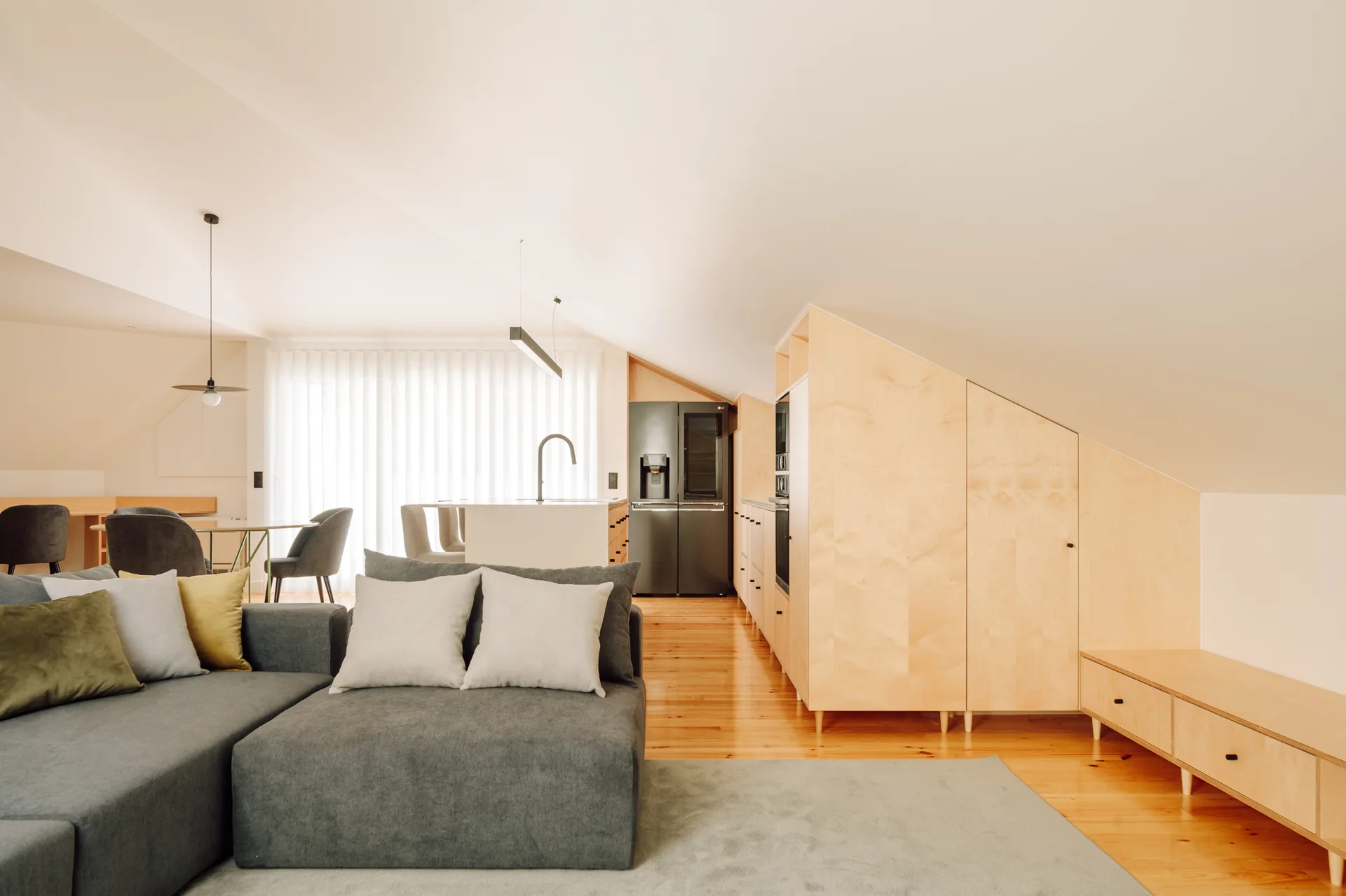 A contemporary open-plan attic living room featuring a grey sectional sofa, light wood floors, and custom cabinets tailored to a sloped ceiling.