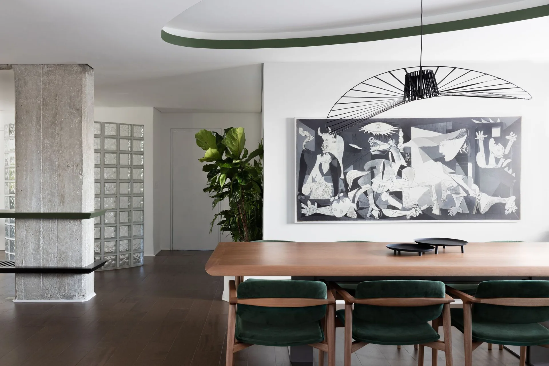A modern dining room features a wooden table with forest green chairs, a raw concrete column, and a large monochromatic cubist art print.