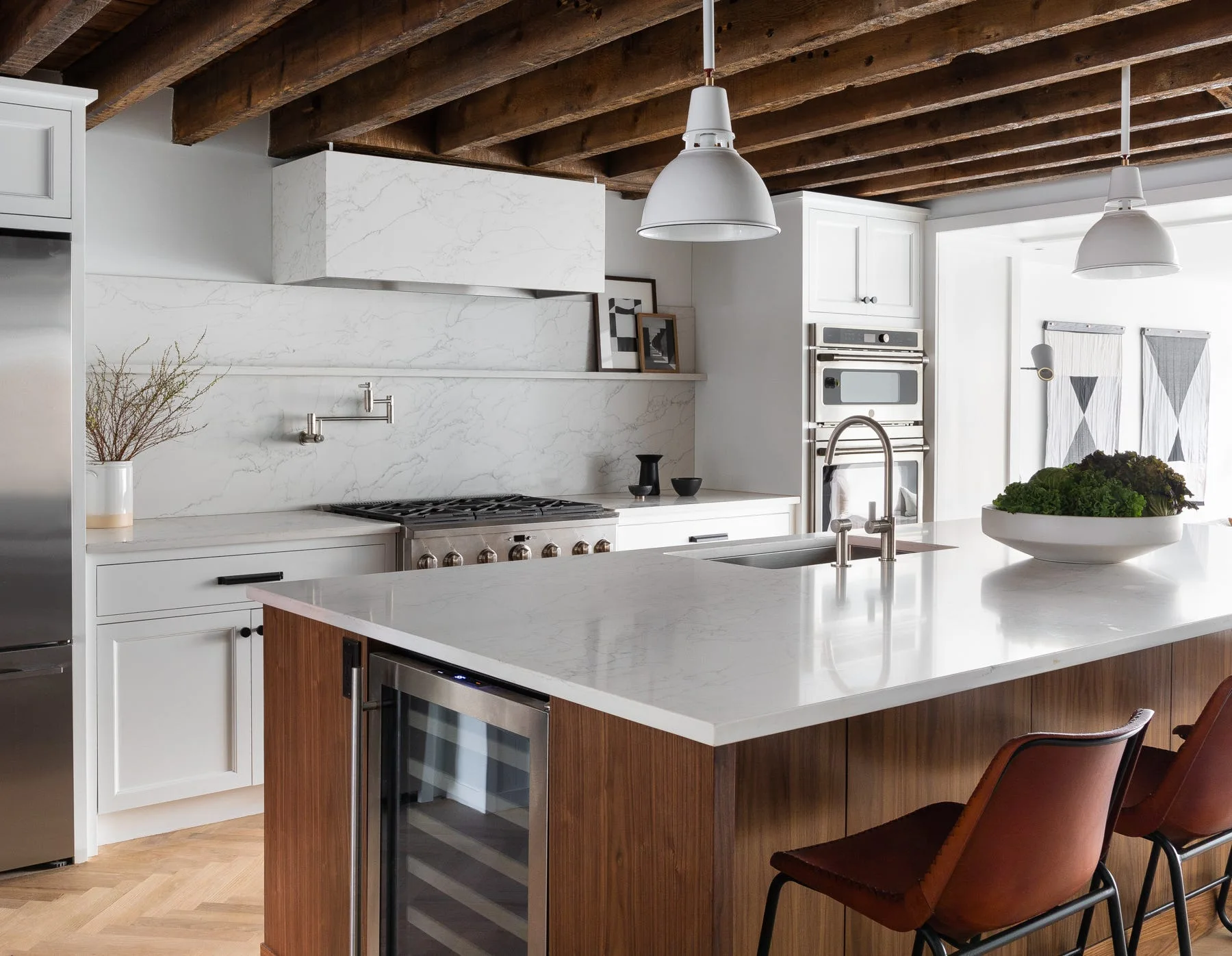 A modern kitchen with white cabinets, a wood island with brown leather stools, marble-look countertops, and exposed dark wood ceiling beams.