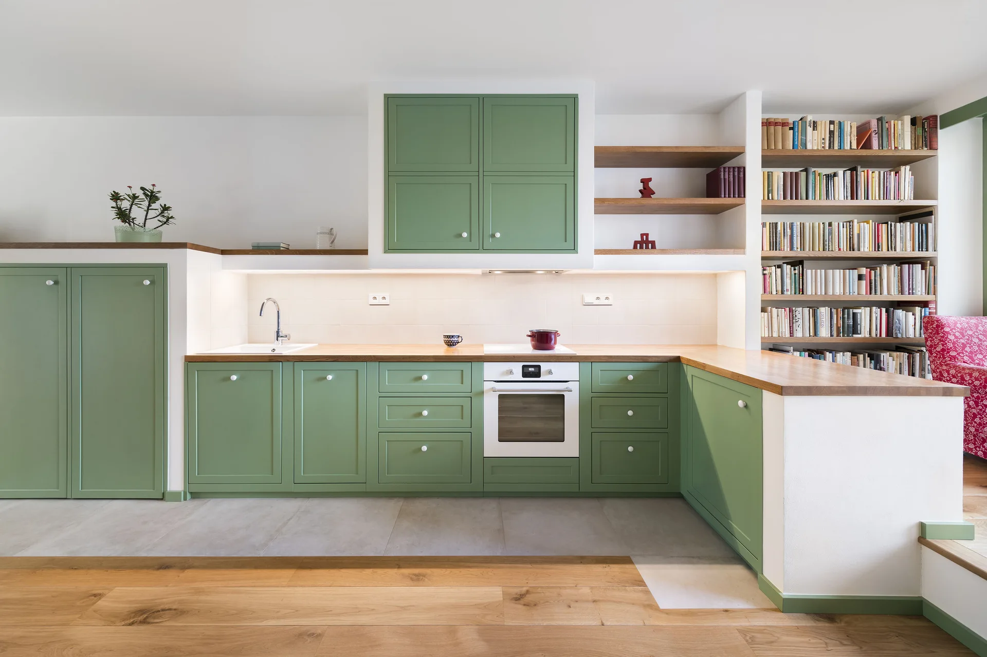 A green shaker kitchen featuring wood countertops, integrated open shelving, a built-in bookshelf filled with books, and grey tiled floors.