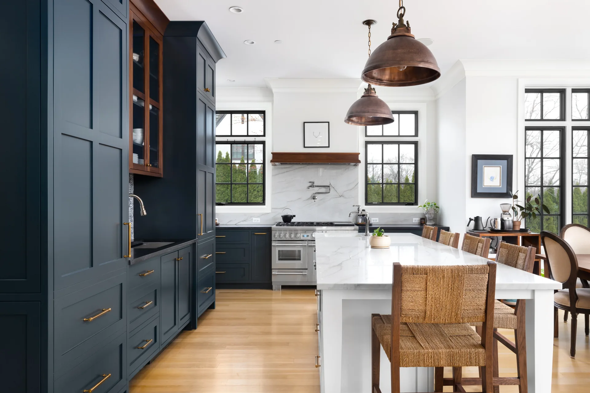 A modern kitchen featuring dark blue cabinetry, a large marble island with woven bar stools, and hanging copper pendant lights.