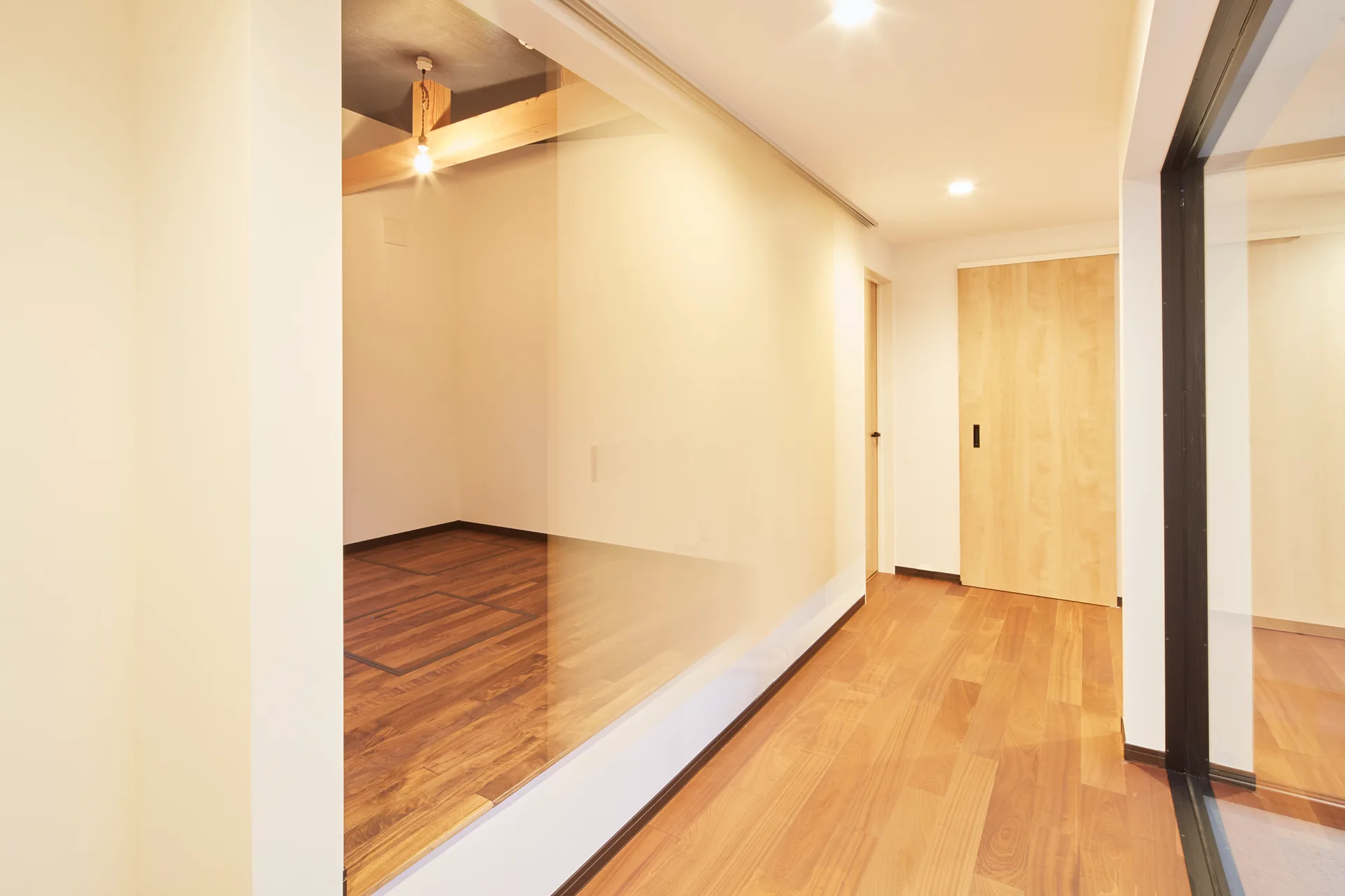 A bright interior hallway features a large glass partition wall, warm hardwood flooring, and a light-toned wood door at the end.