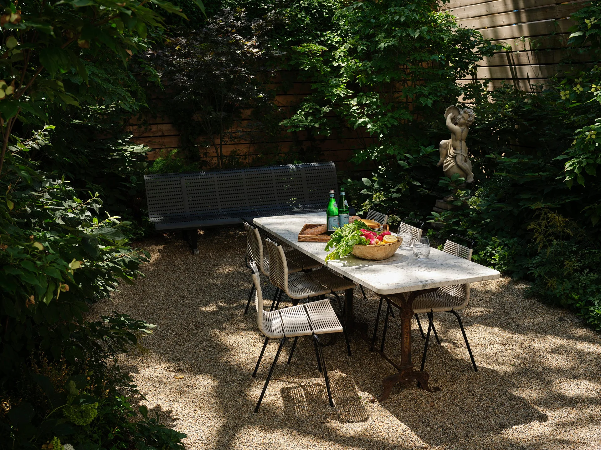 A marble-topped dining table and woven chairs sit on a gravel patio surrounded by dense garden greenery.