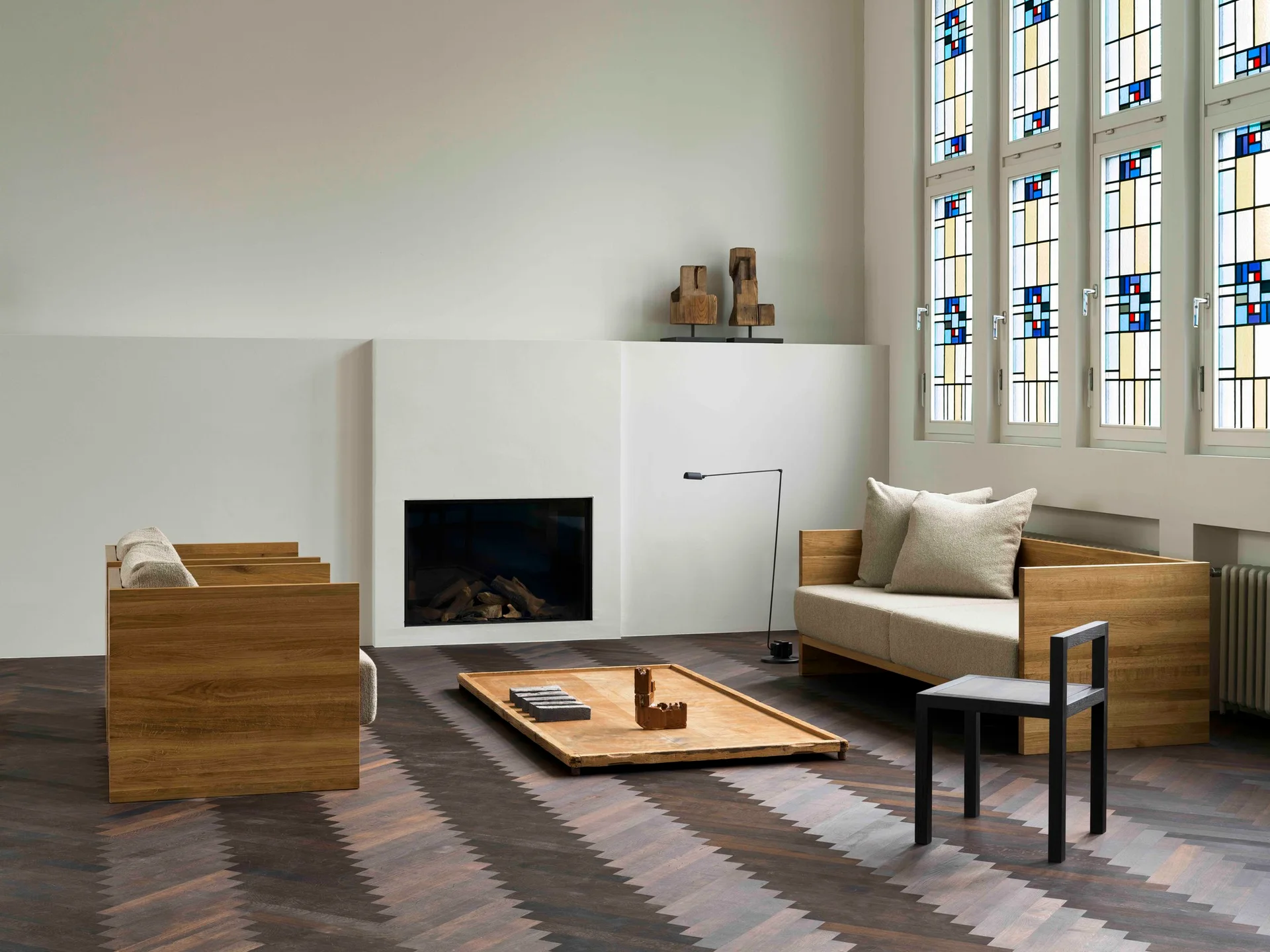 A minimalist living room features wooden sofas and a low table on dark herringbone wood floors near a white fireplace and stained glass windows.