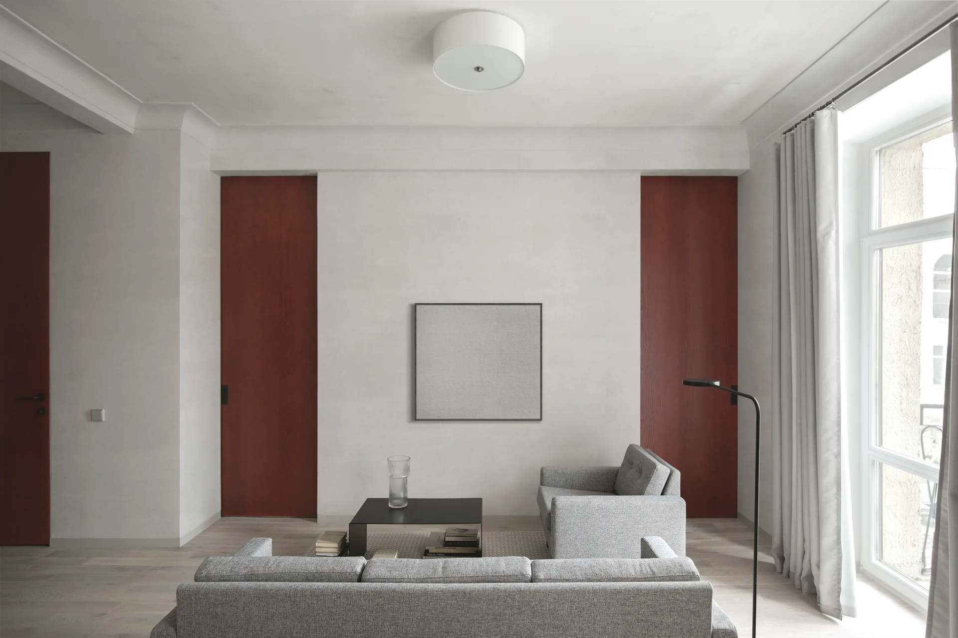 A minimalist living room featuring grey upholstered furniture, a black coffee table, and two tall reddish-brown wood doors framing central artwork.