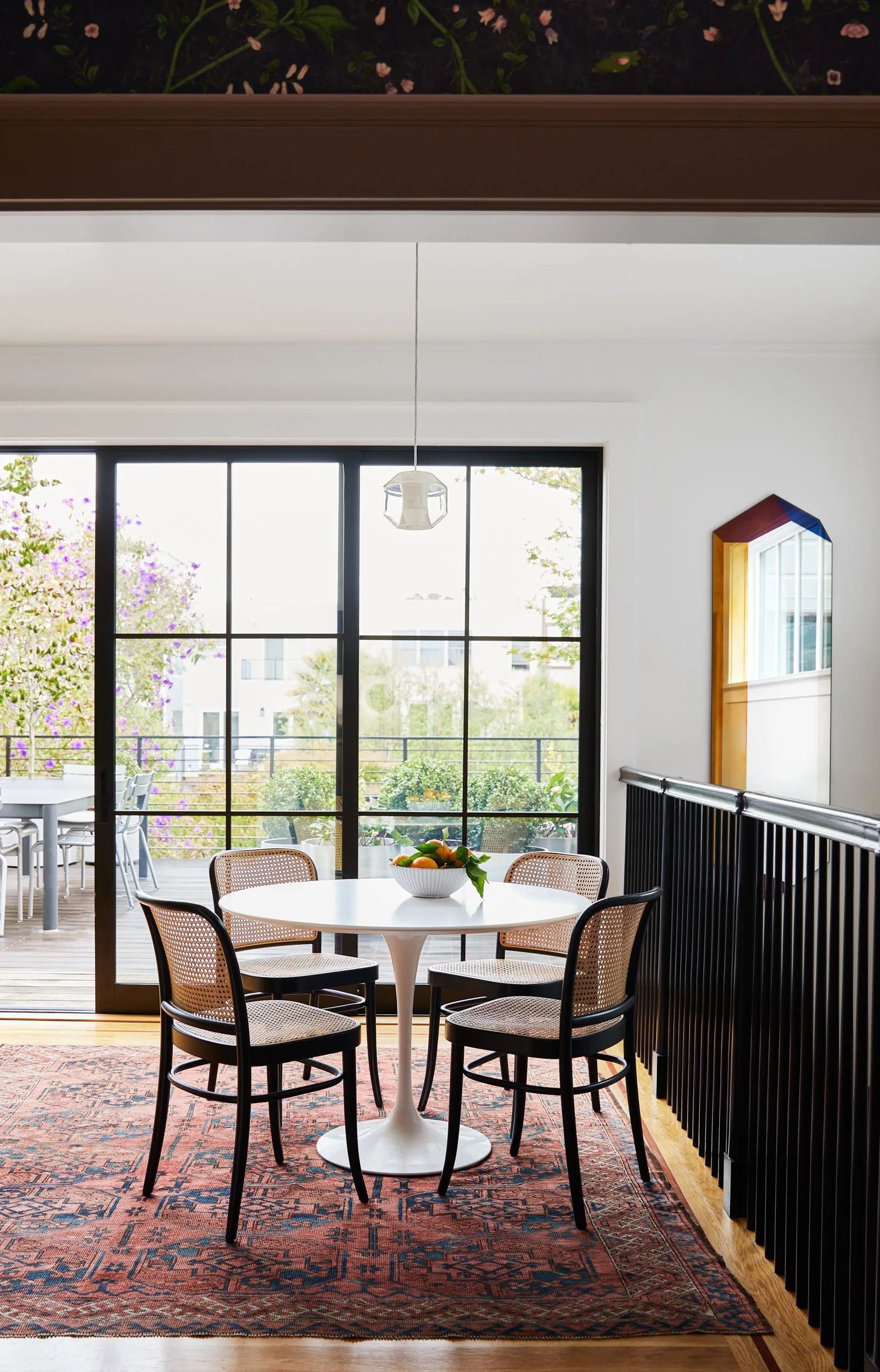 A white round tulip table and four black cane-back chairs sit on a red patterned rug in front of large black-framed glass sliding doors.