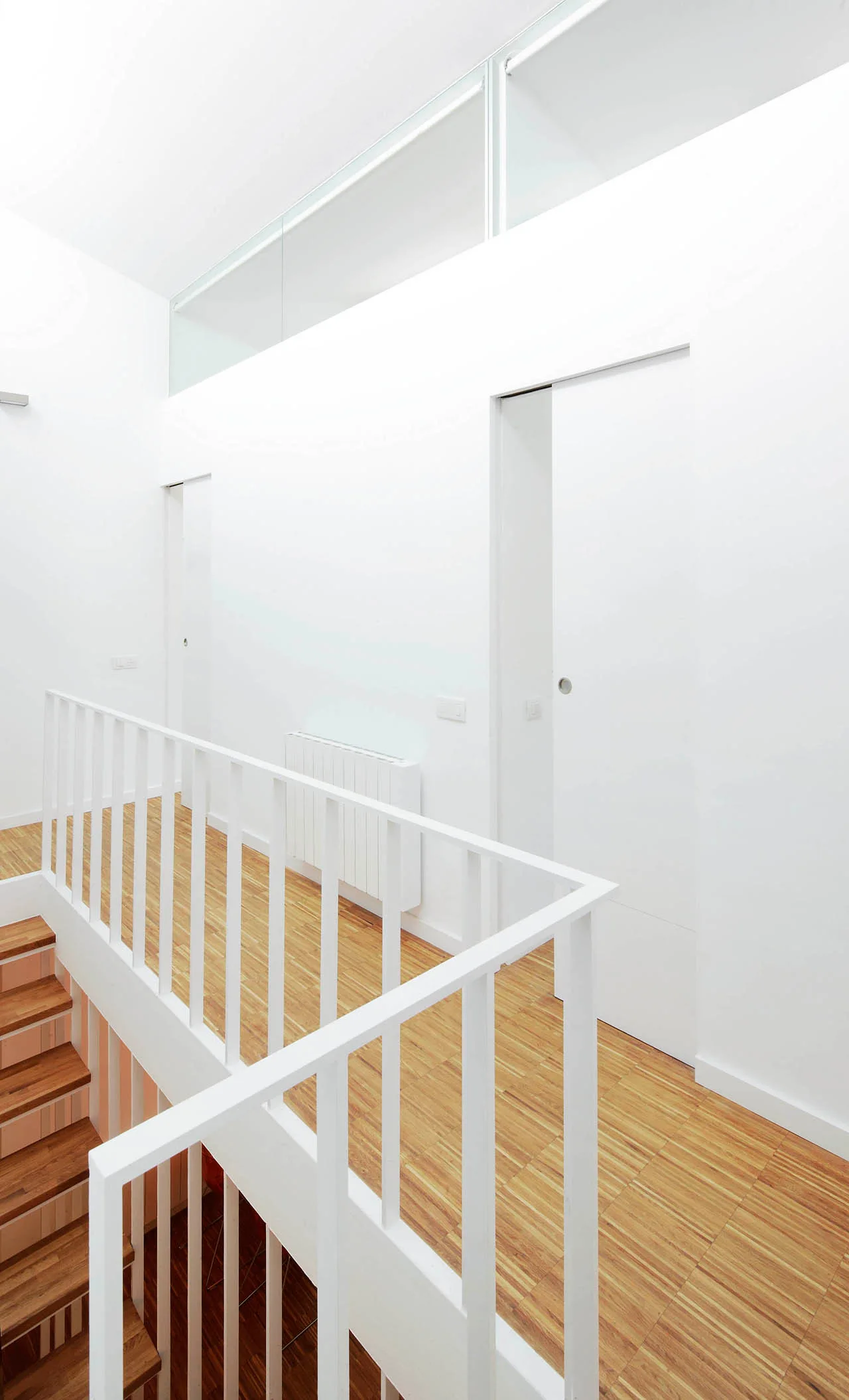 A bright minimalist hallway landing featuring light wood flooring, white sliding doors, and a white metal staircase railing.