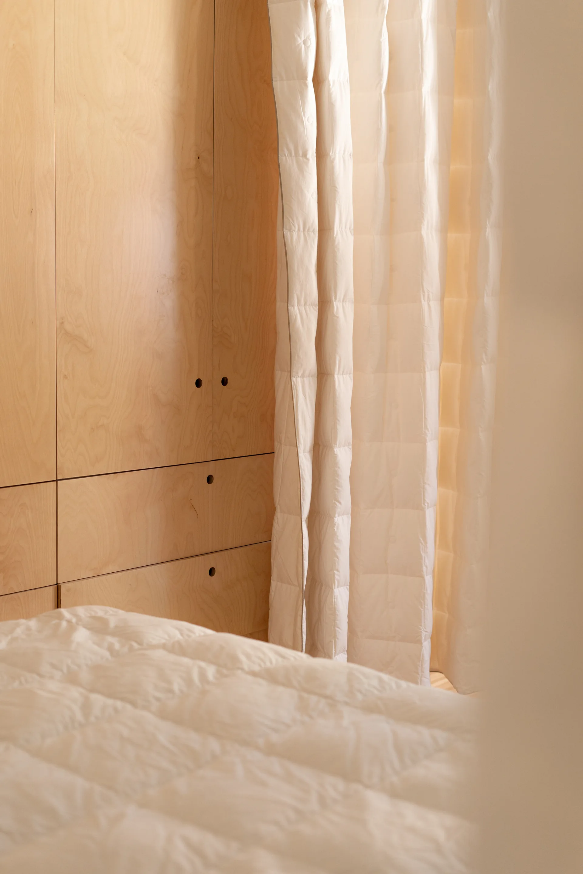 A minimalist bedroom featuring flat panel light wood wardrobe doors with circular pulls next to a white quilted curtain and matching bedding.
