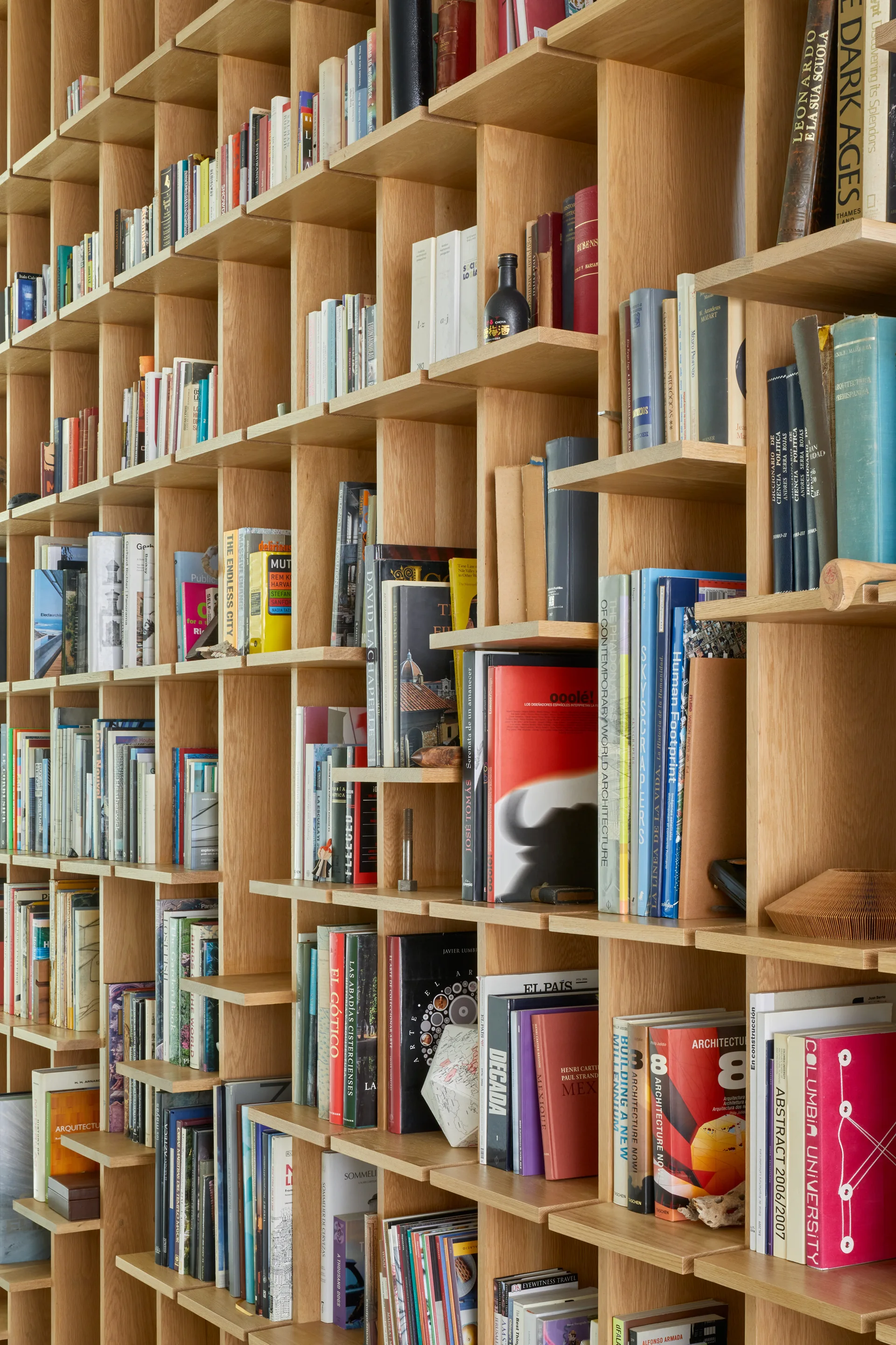 A floor-to-ceiling light wood bookshelf with a staggered grid design is packed with colorful books and small decorative items.