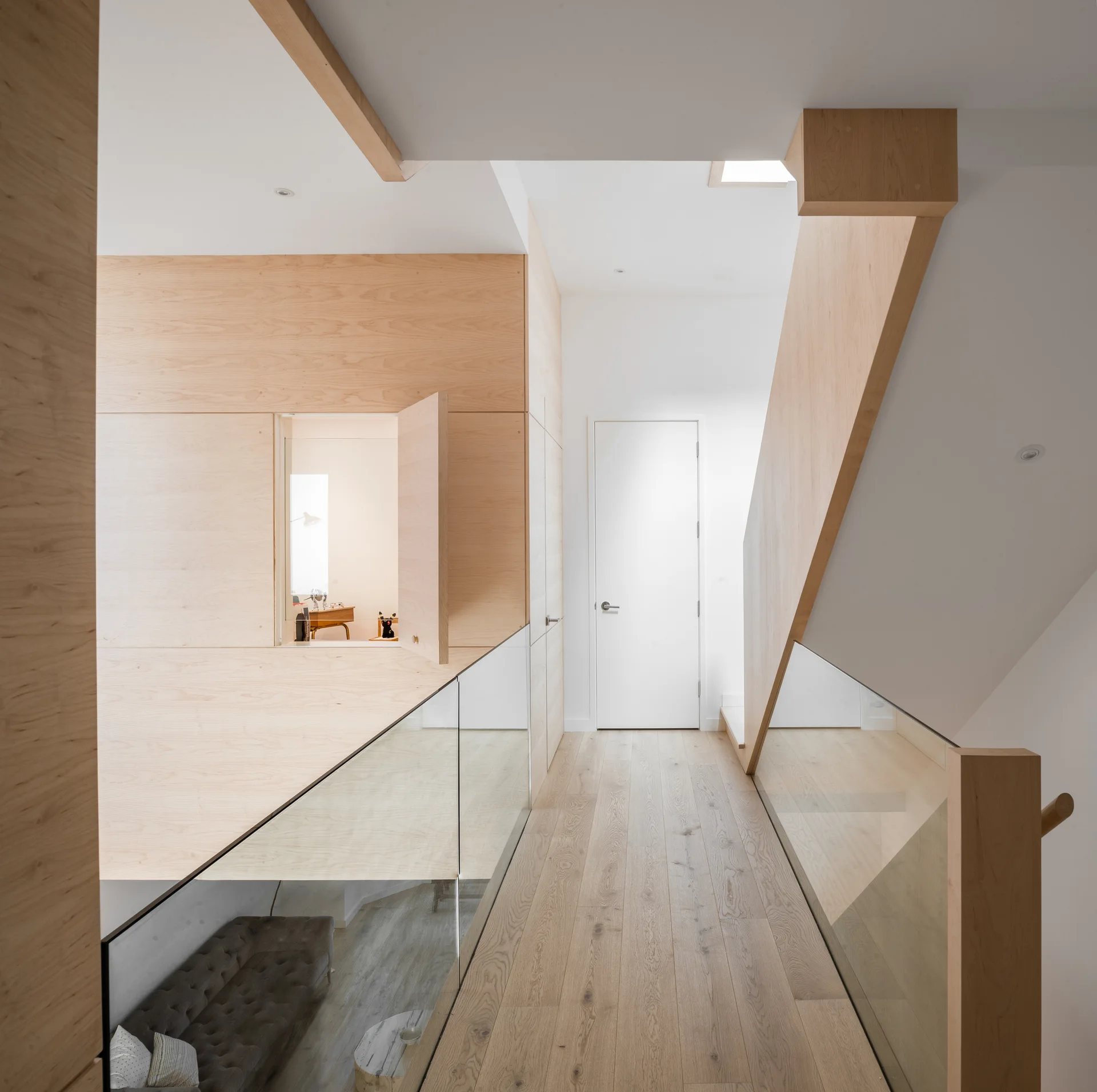 A modern residential hallway landing features light wood wall paneling with a flush door, matching oak flooring, and a glass-paneled staircase railing.