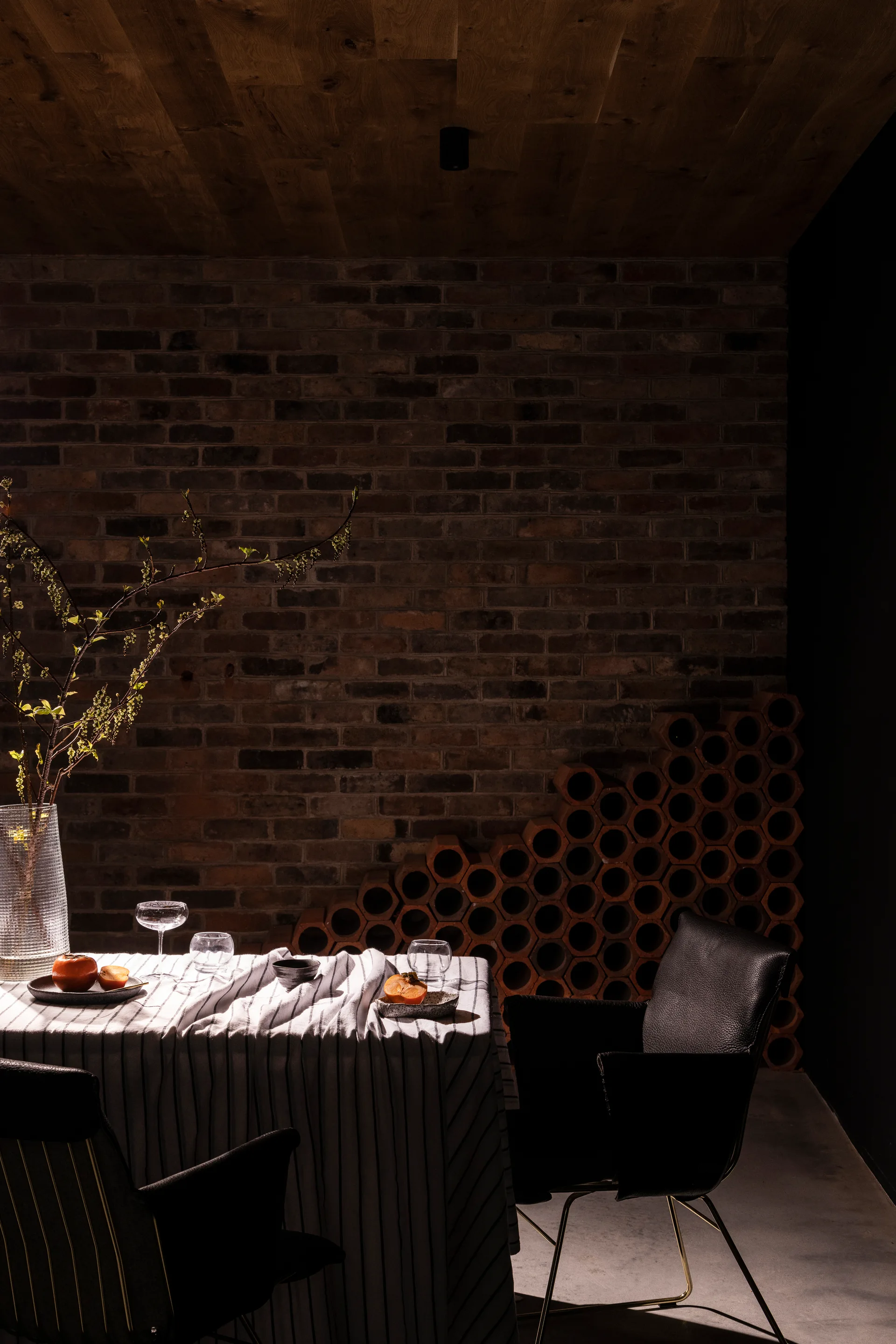 A dimly lit dining area features a table with a striped tablecloth, black chairs, an exposed brick wall, and stacked terracotta wine racks.