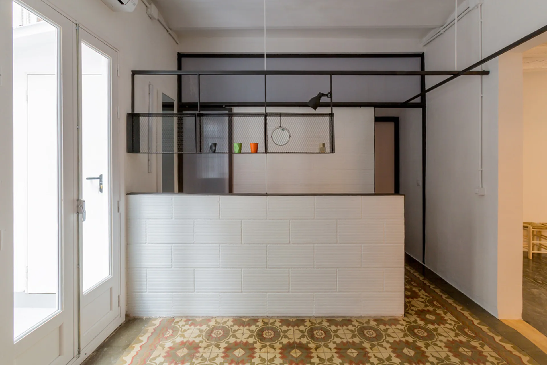 An interior space featuring a low white brick partition wall beneath a suspended black metal mesh shelving unit and patterned floor tiles.