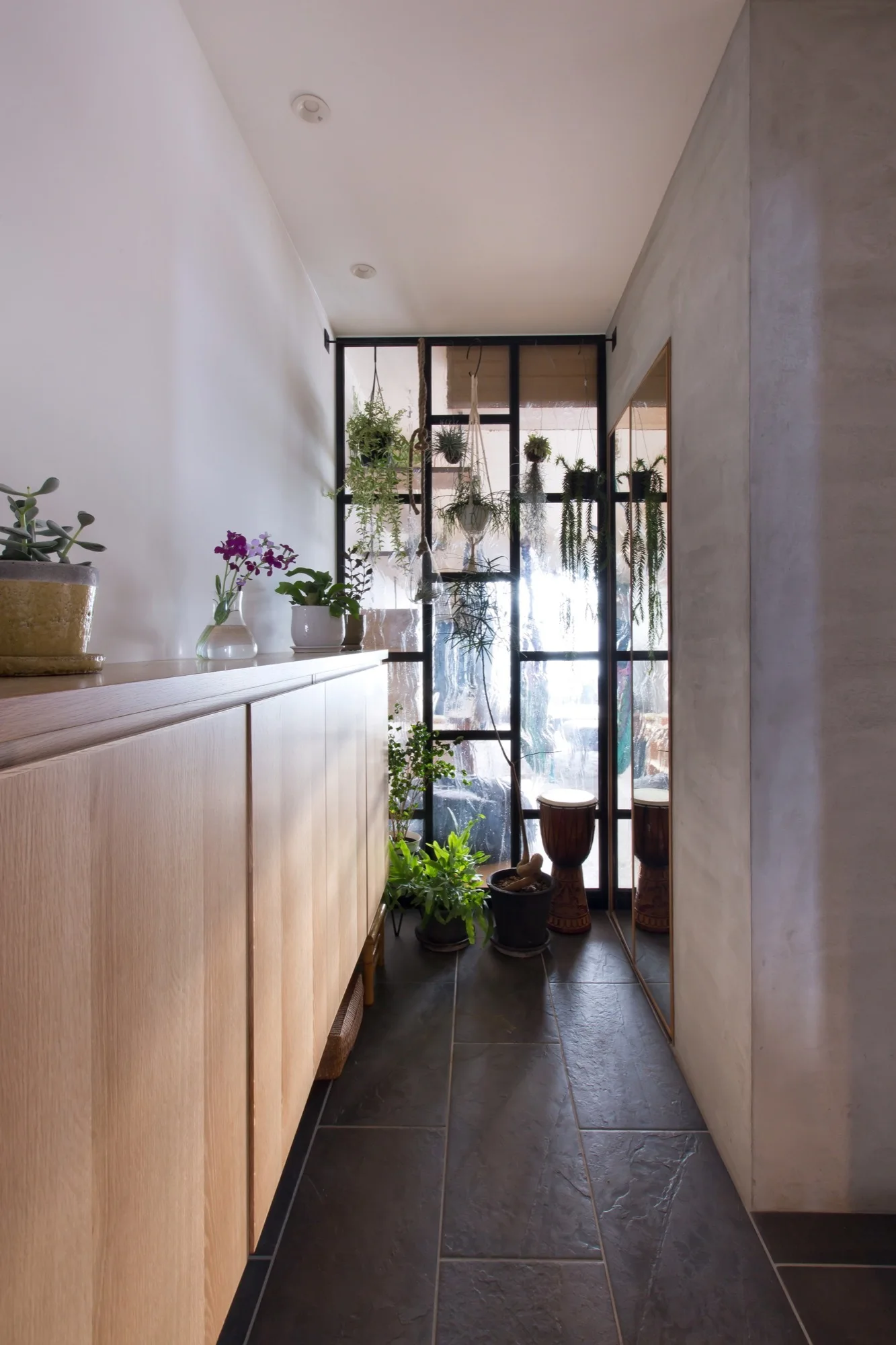 A narrow hallway with a light wood storage cabinet, dark stone floor tiles, and a black metal-framed glass partition filled with hanging plants.