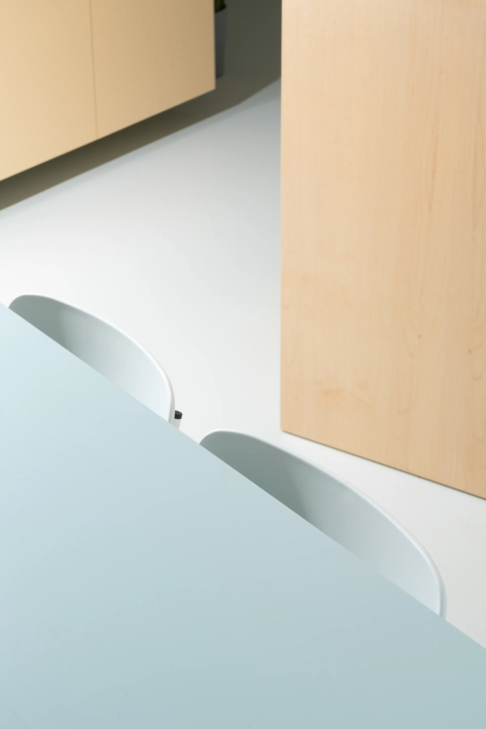 A high-angle close-up of a matte light blue dining table with two light grey chairs set against minimal light wood cabinets and a smooth white floor.