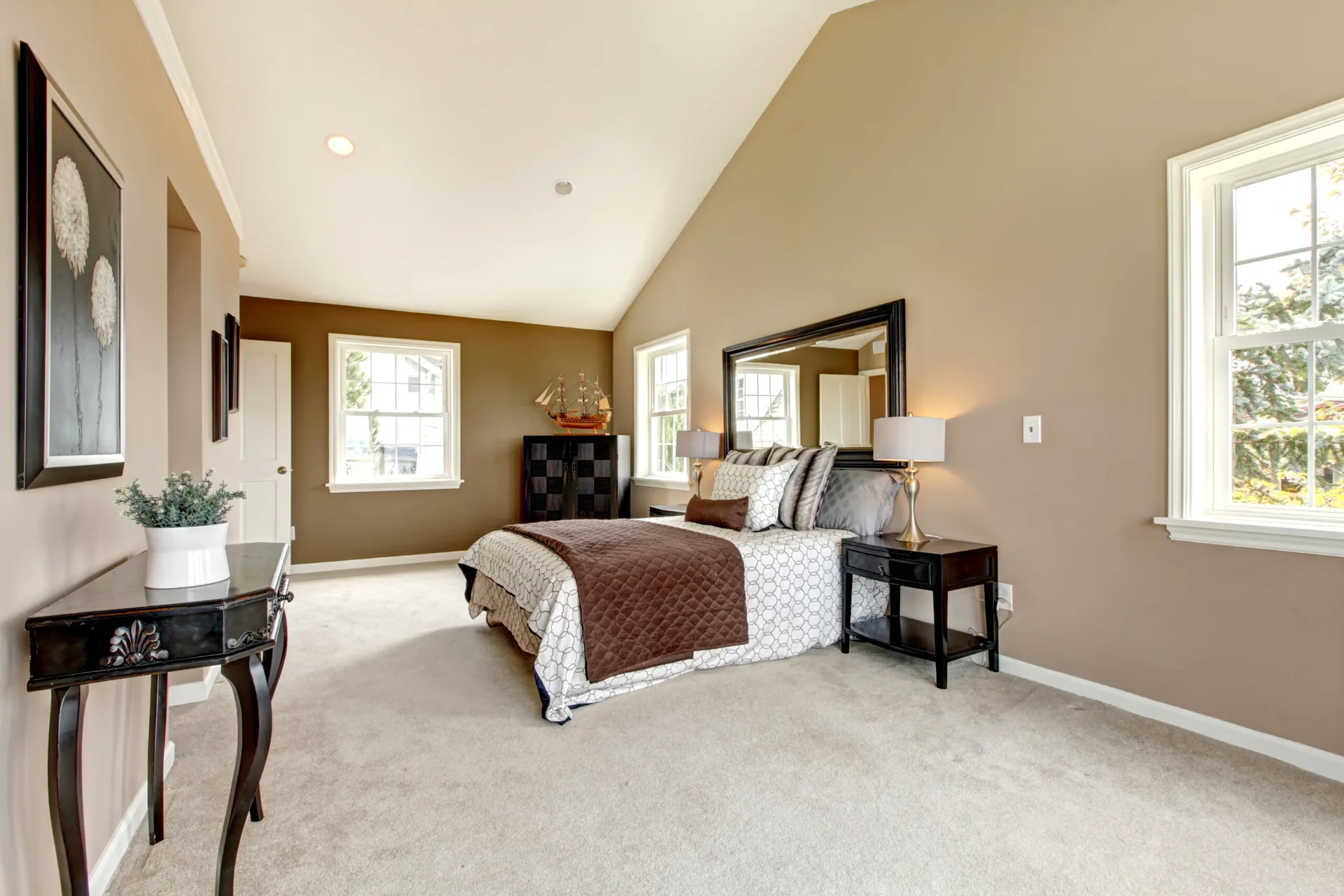 A carpeted bedroom with a vaulted ceiling, a bed flanked by black nightstands, a large mirror on the wall, and multiple windows.