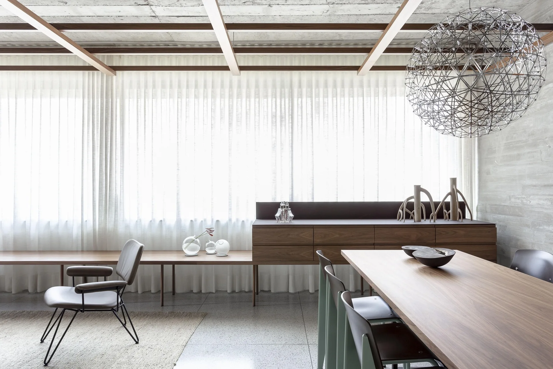 A modern dining room features a wooden dining table, a low sideboard against floor-to-ceiling sheer white curtains, and an exposed concrete wall.