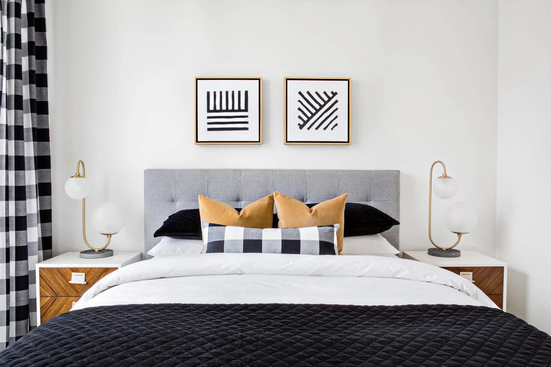 A modern bedroom features a grey tufted headboard, two geometric framed prints, wood nightstands with brass sphere lamps, and yellow and plaid pillows.