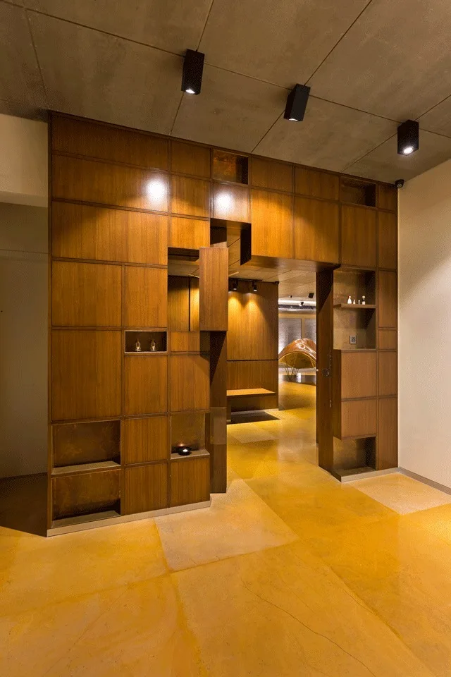 A large warm wood room divider with built-in shelving niches stands on a yellow floor beneath a concrete ceiling with black spotlights.