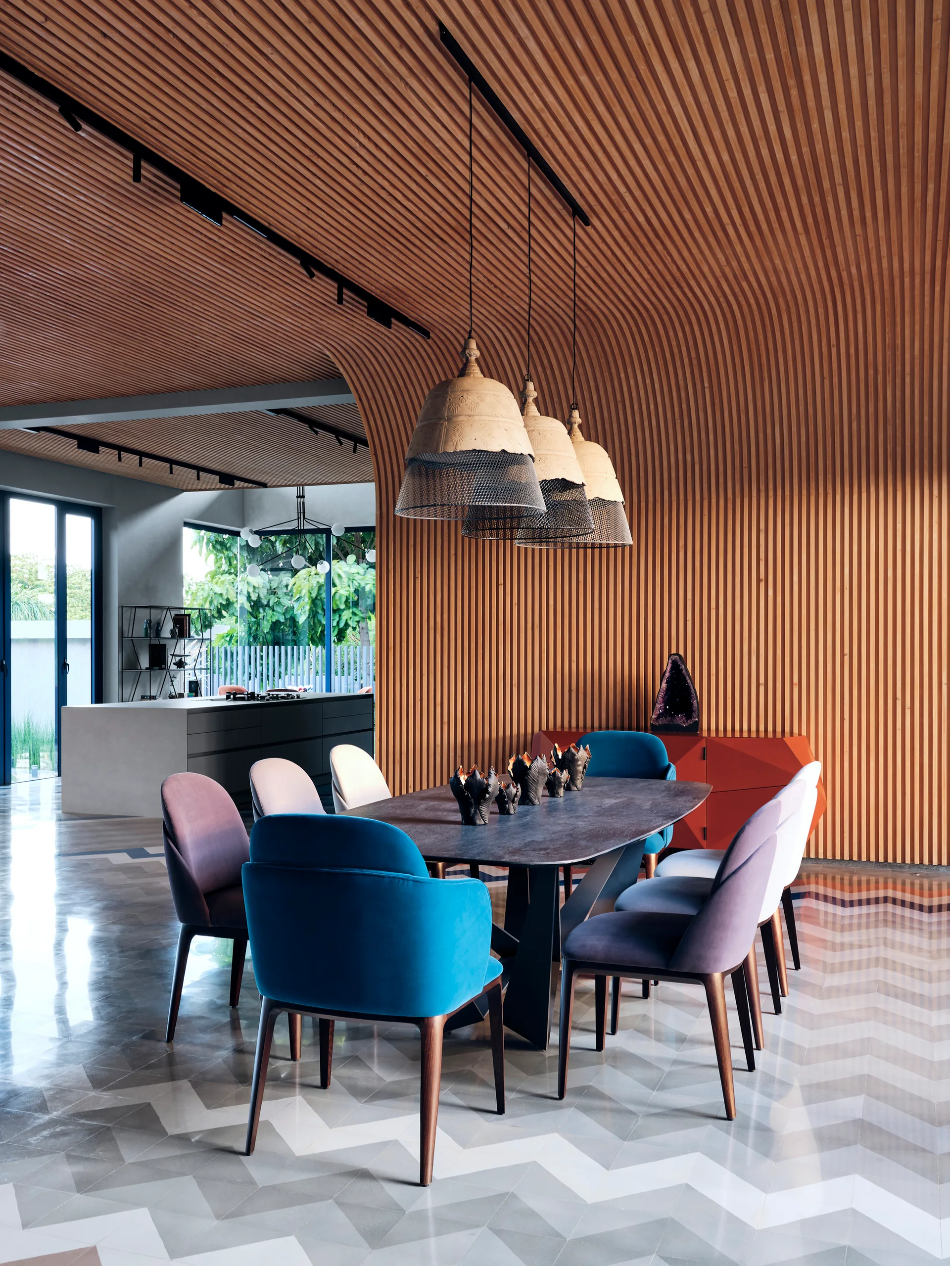 A modern dining room featuring a curved wood slat wall and ceiling, a dark dining table with colorful chairs, and three large pendant lights.