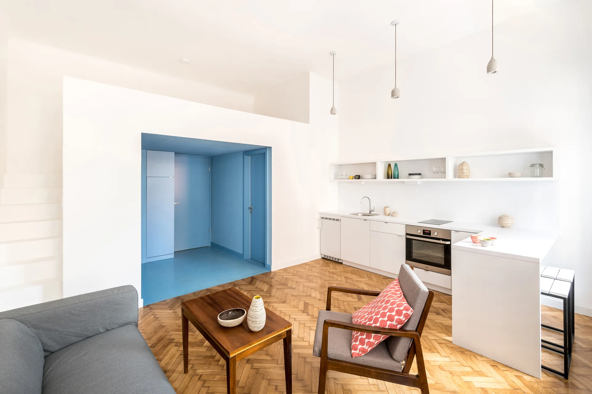 A bright living room and kitchenette featuring wooden herringbone flooring, white cabinets, mid-century furniture, and a bold blue recessed entryway.