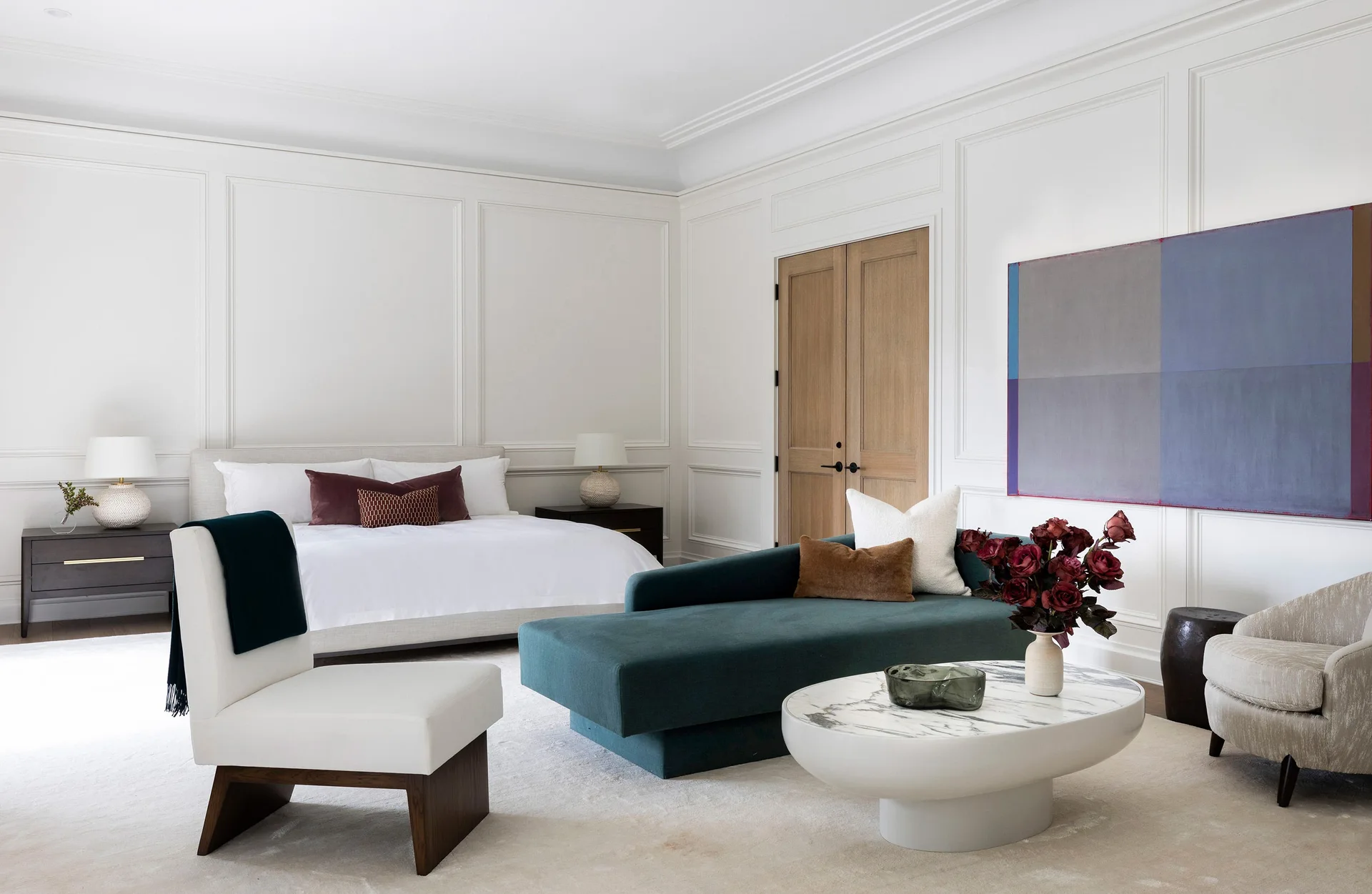 A white bedroom features paneled walls, a bed with white linens, and a deep teal chaise lounge paired with a marble-topped coffee table.