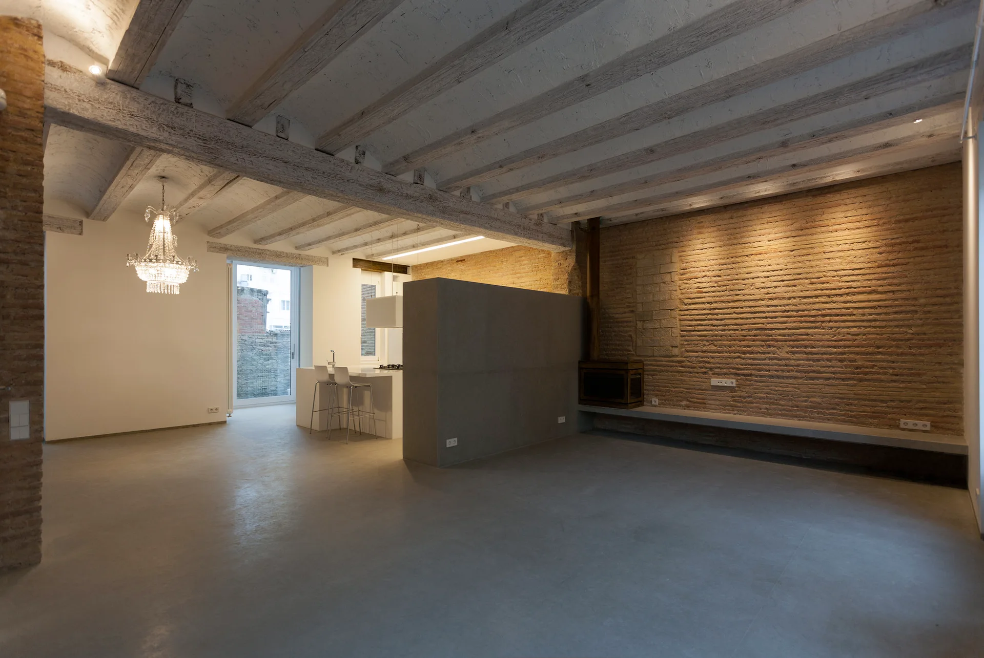 An open-concept living room features an exposed brick wall, a white wooden beamed ceiling, and a grey partition wall adjacent to a white kitchen island.