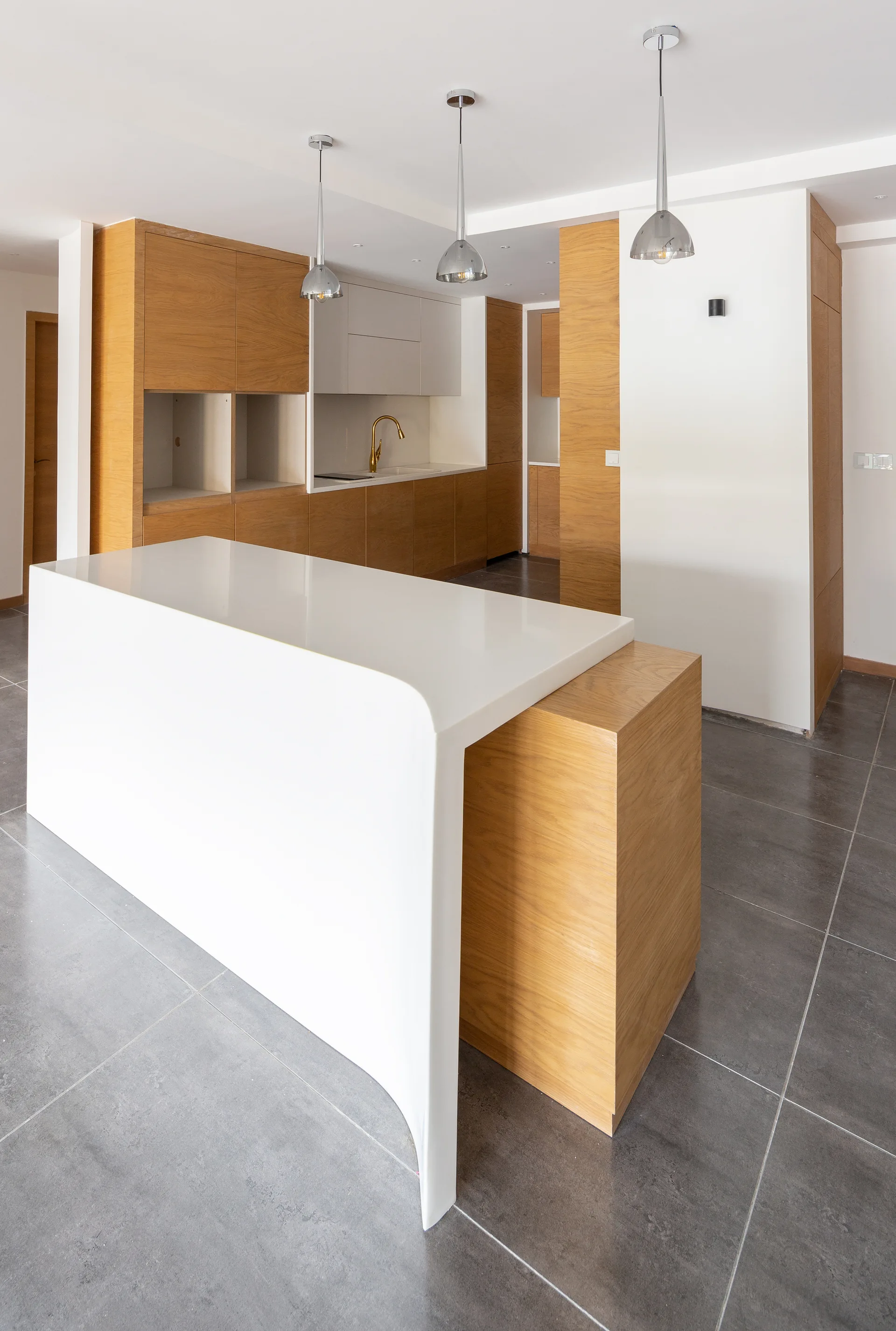 A modern kitchen featuring a curved white waterfall island nested with oak wood, matching oak cabinets, and grey tiled floors.