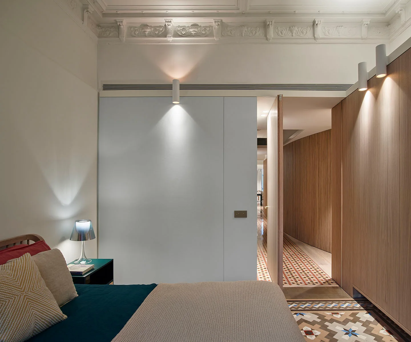 A bedroom features ornate white ceiling moldings, a modern wall partition, and a view into a hallway with colorful geometric floor tiles.