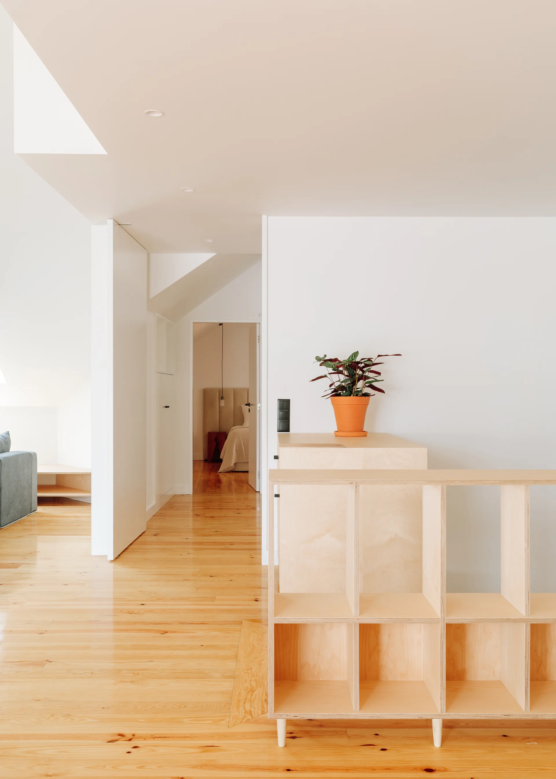 A bright residential hallway with warm polished wood flooring, light plywood modular shelving, and a bedroom visible in the background.