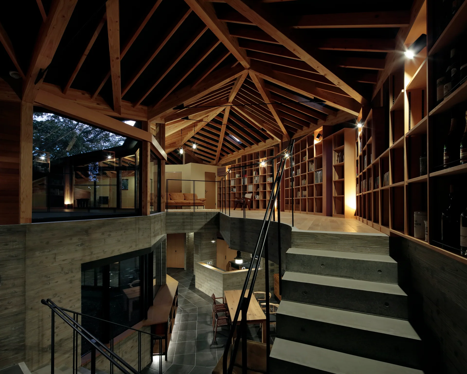 A split-level home interior features a raw concrete staircase, a mezzanine lined with floor-to-ceiling wooden bookshelves, and an exposed timber ceiling.