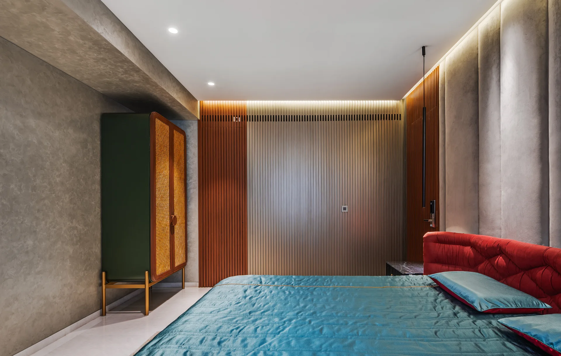 A modern bedroom features a teal quilted bed with a red headboard next to a green cane cabinet and vertical wood slatted walls.
