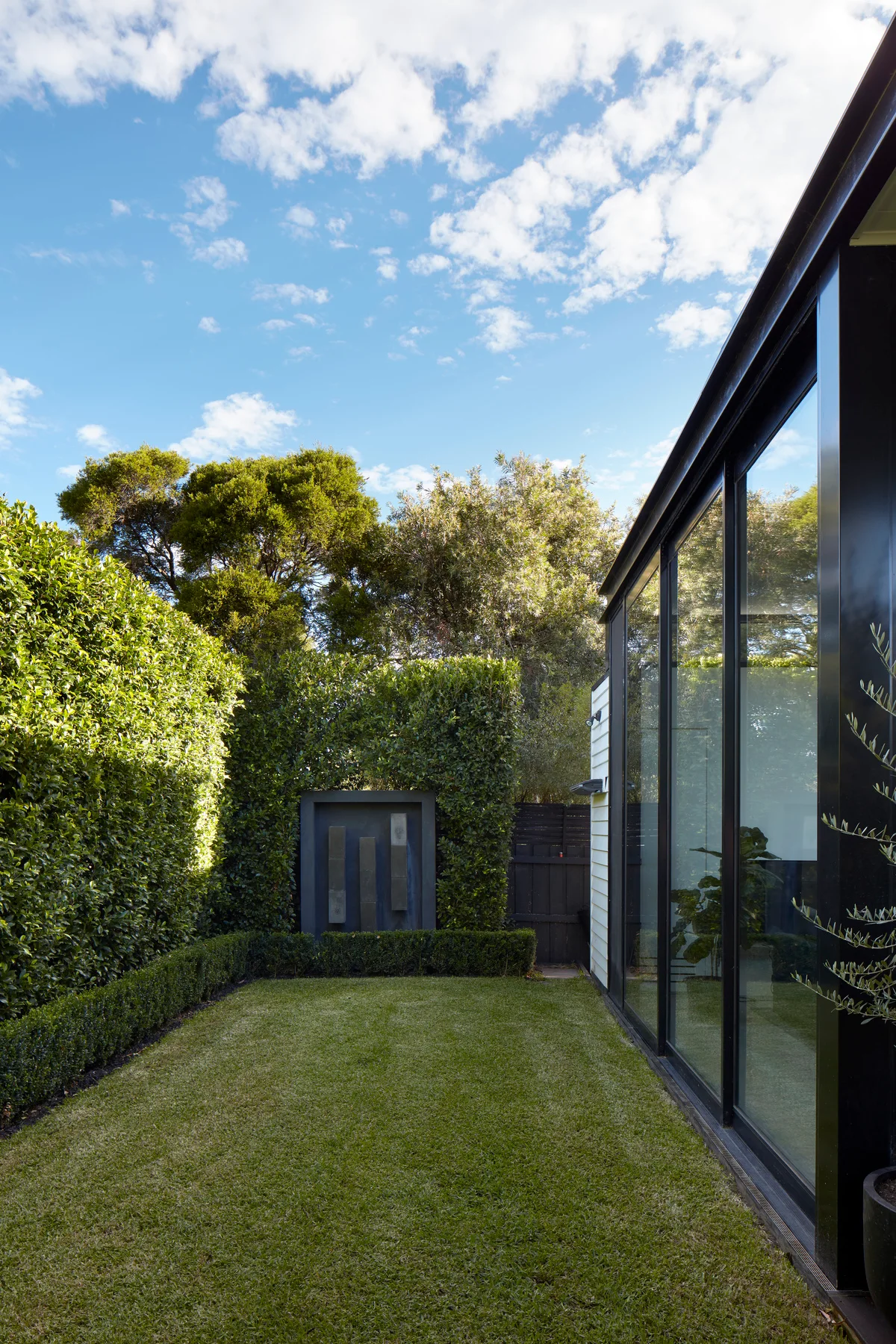 A modern residential garden features a mowed green lawn surrounded by tall, manicured hedges, a minimalist sculpture, and a large black-framed glass wall.