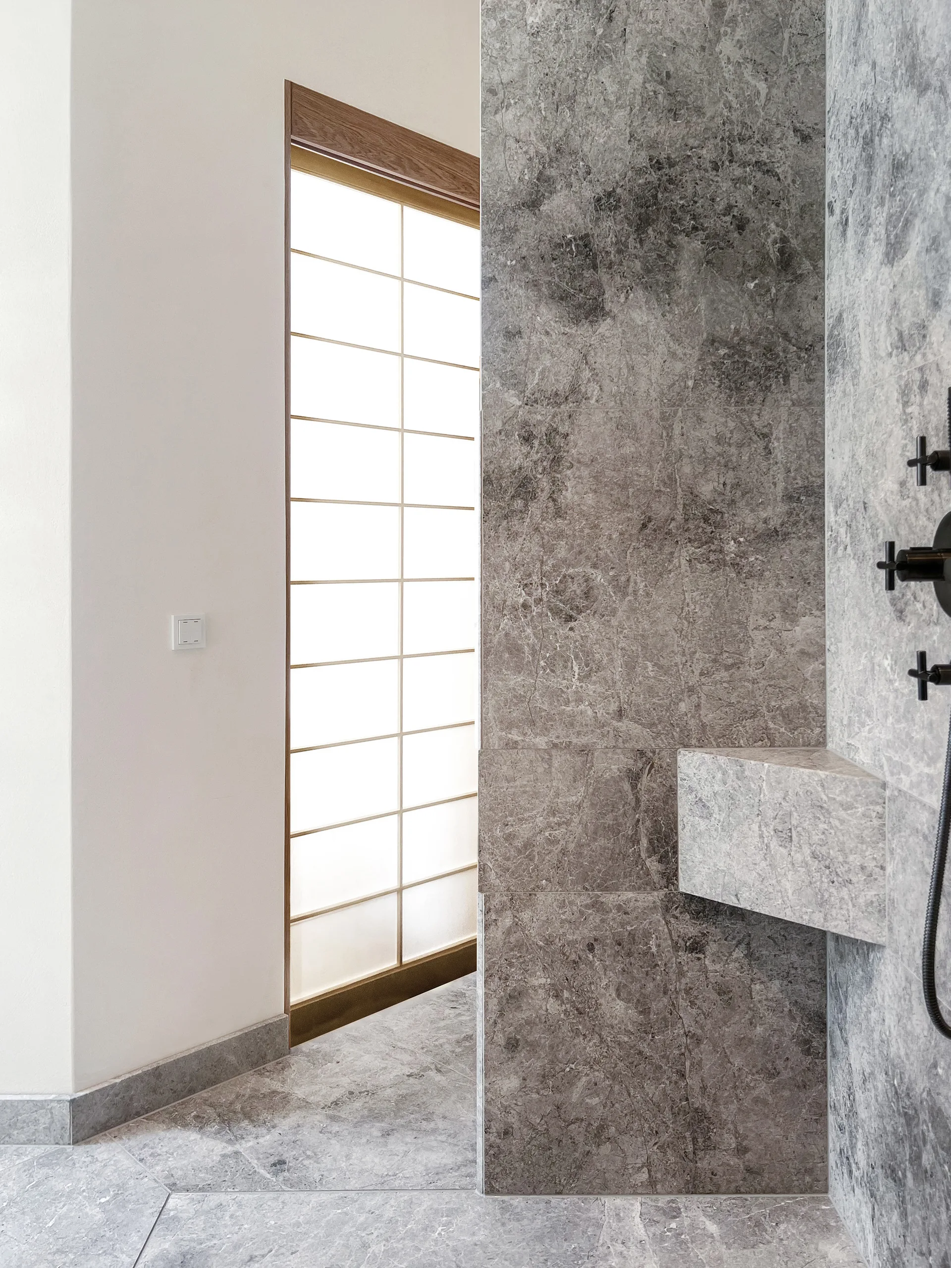 A minimalist bathroom shower features grey veined marble walls, a matching integrated stone corner shelf, and a grid-patterned translucent screen.