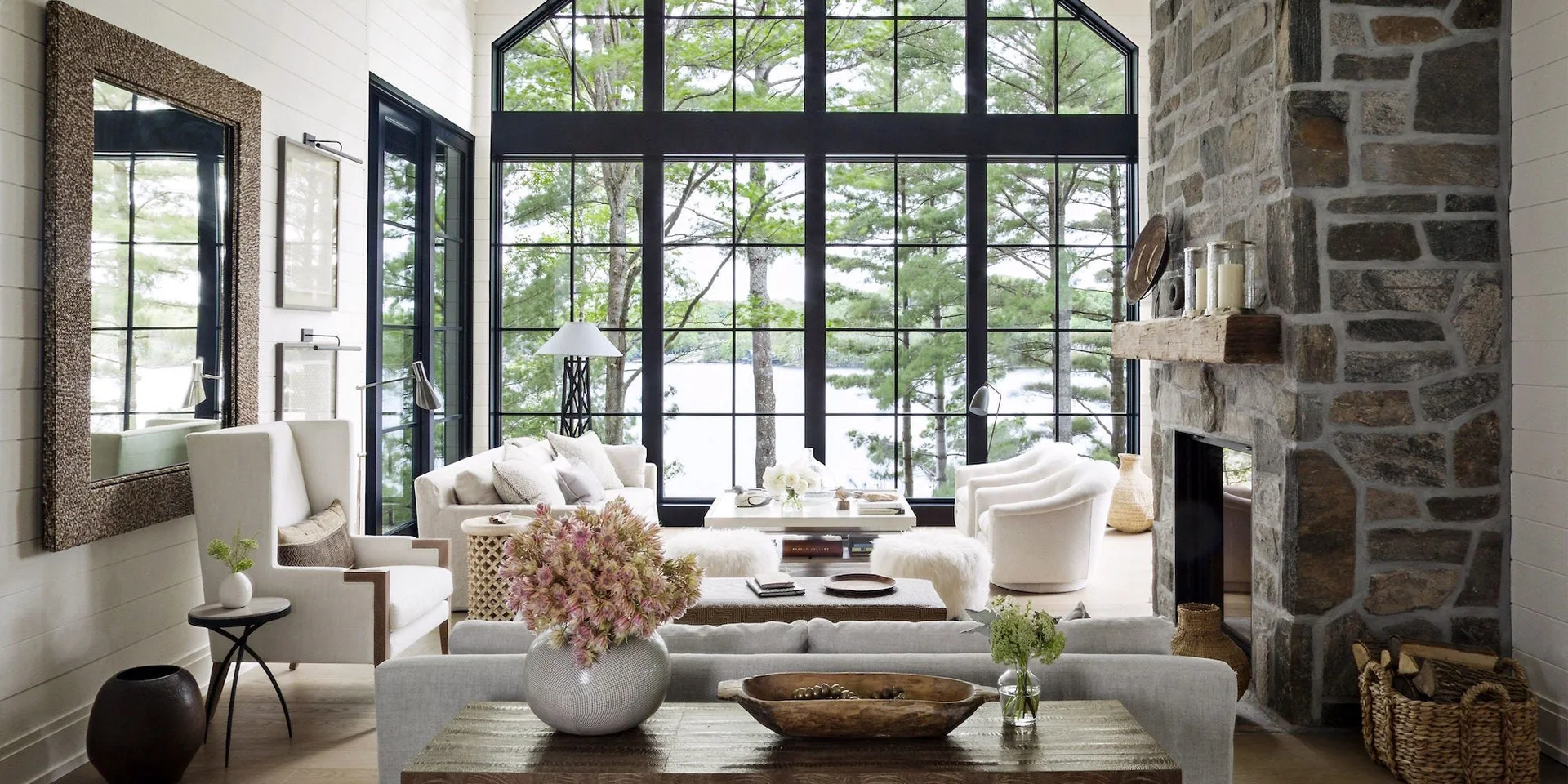 A bright living room features cream sofas, a massive stone fireplace with a wood mantel, and tall black-framed arched windows showing trees.
