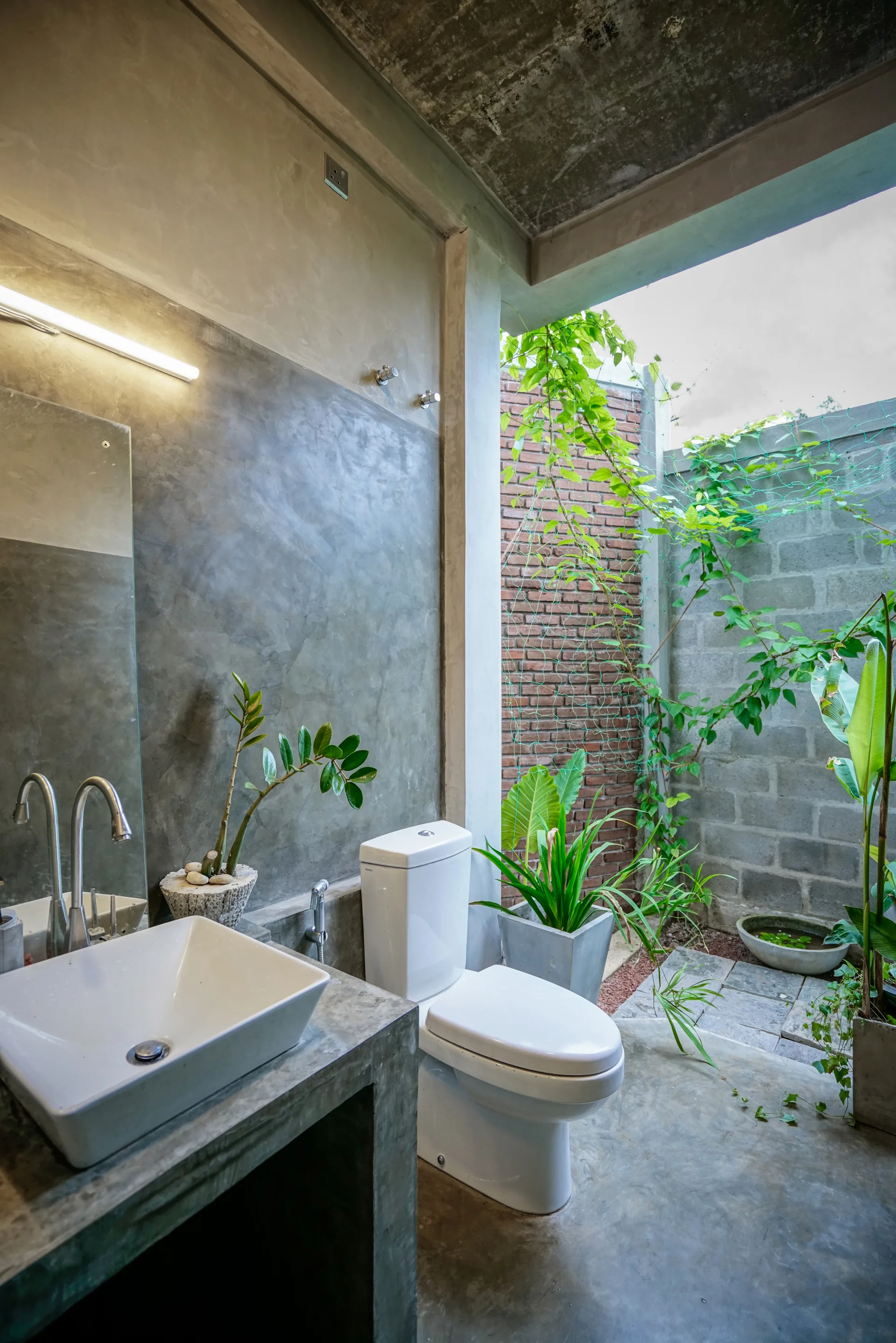 A minimalist concrete bathroom features a white vessel sink on a concrete vanity and opens directly onto an outdoor garden courtyard.